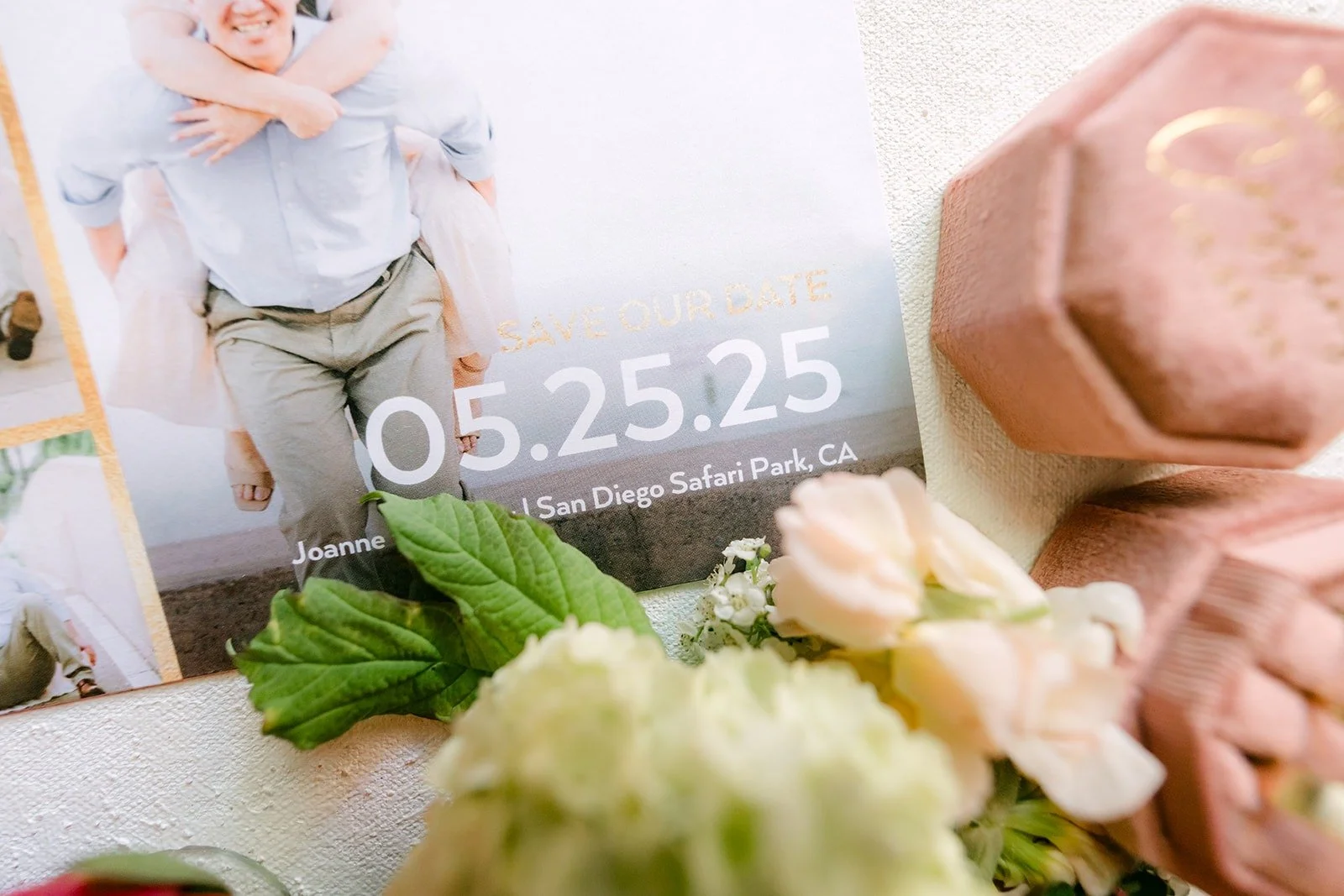 A photo of the couple with a 05.25.25 date card, pink ring boxes and white flowers