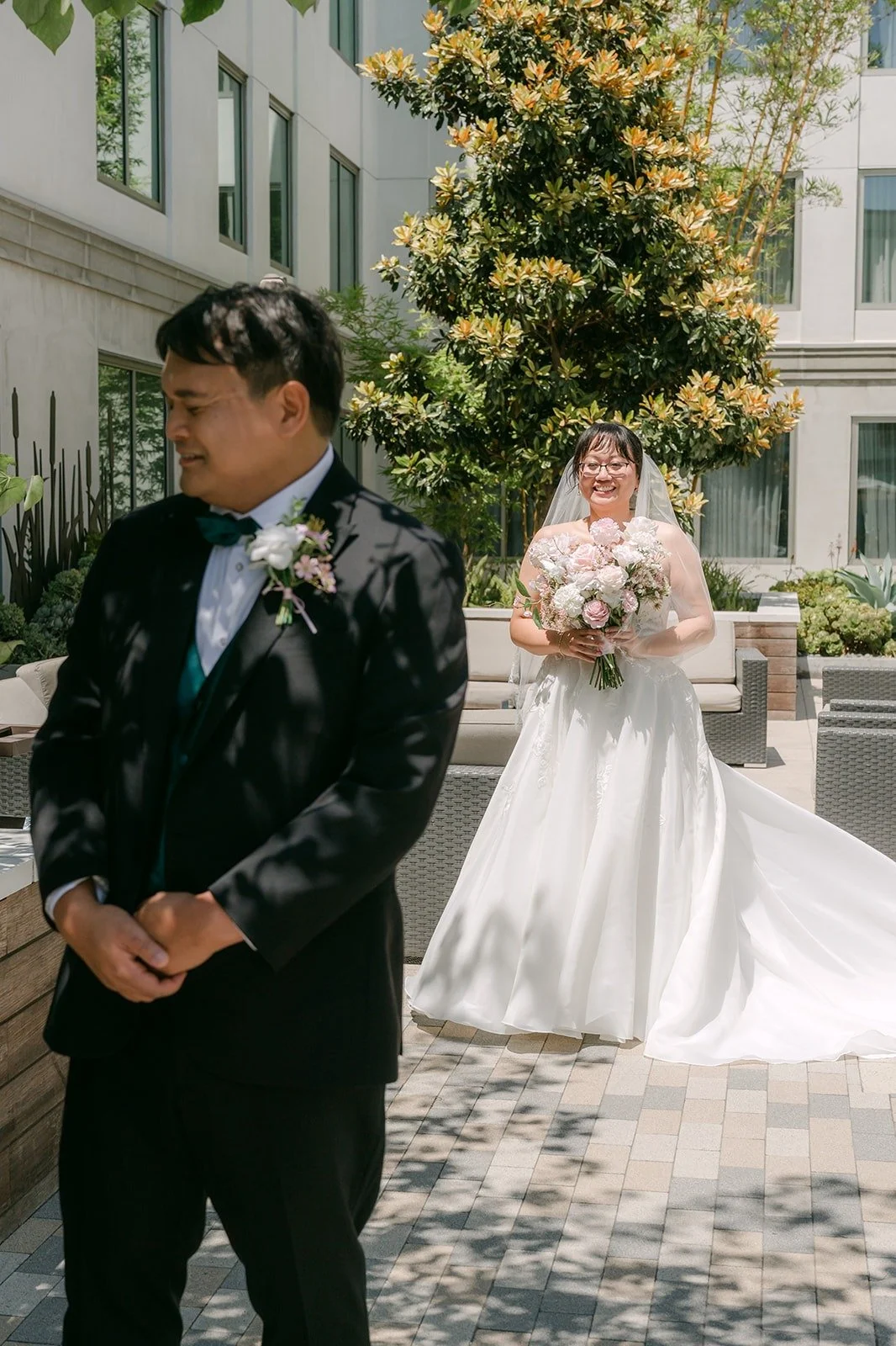Stella walking up behind James on the courtyard lawn with her bouquet