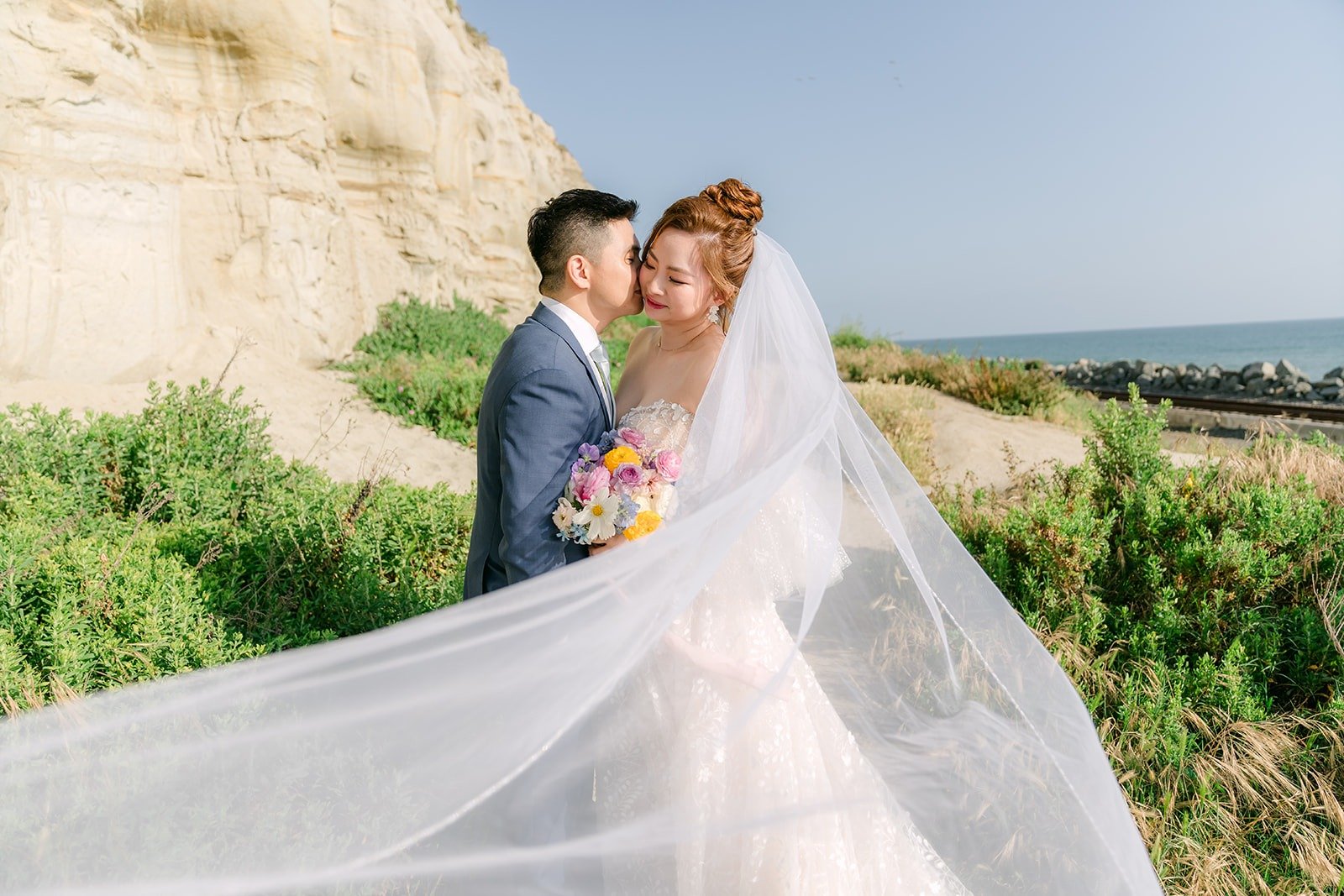 Joanna's long veil catching the wind at the beach as Jerrick holds her