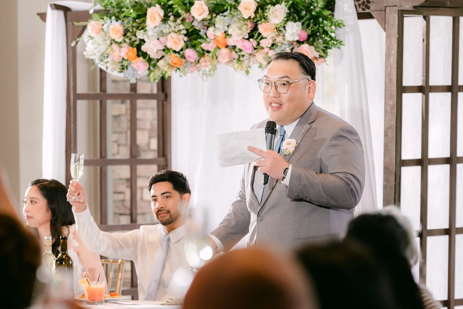 A groomsman speaking into the microphone as Natalie and Mark laugh