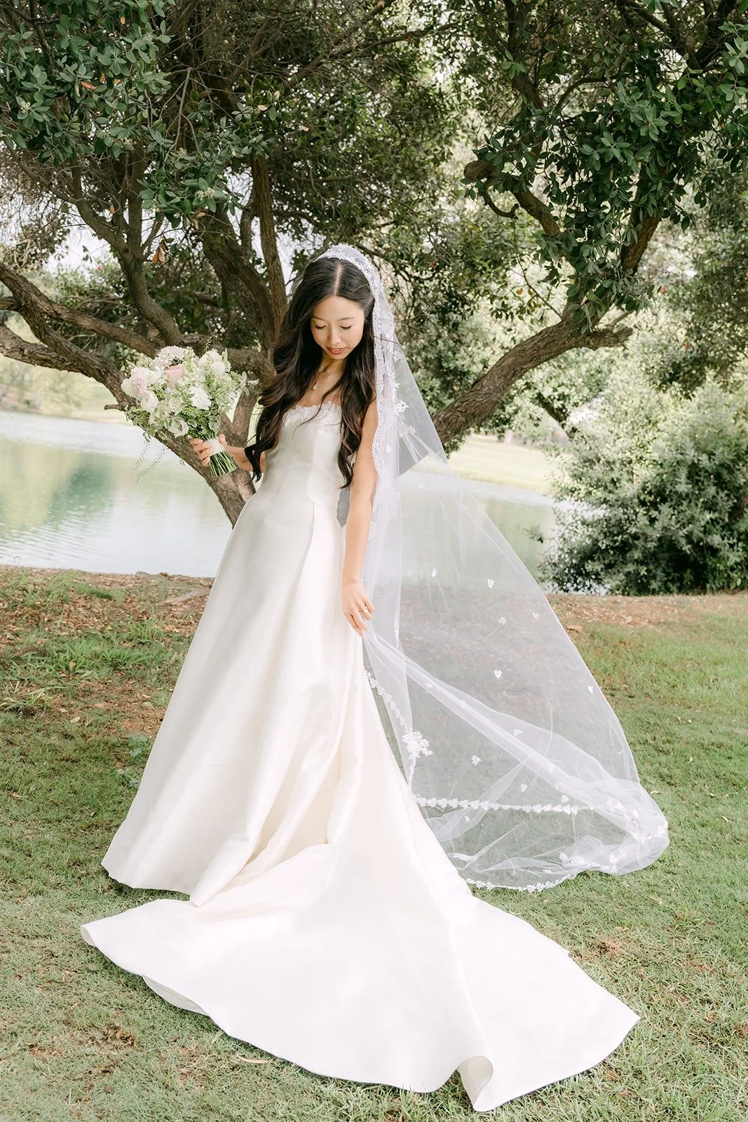 Kristine in her gown with her cathedral veil spread across the grass beside the lake