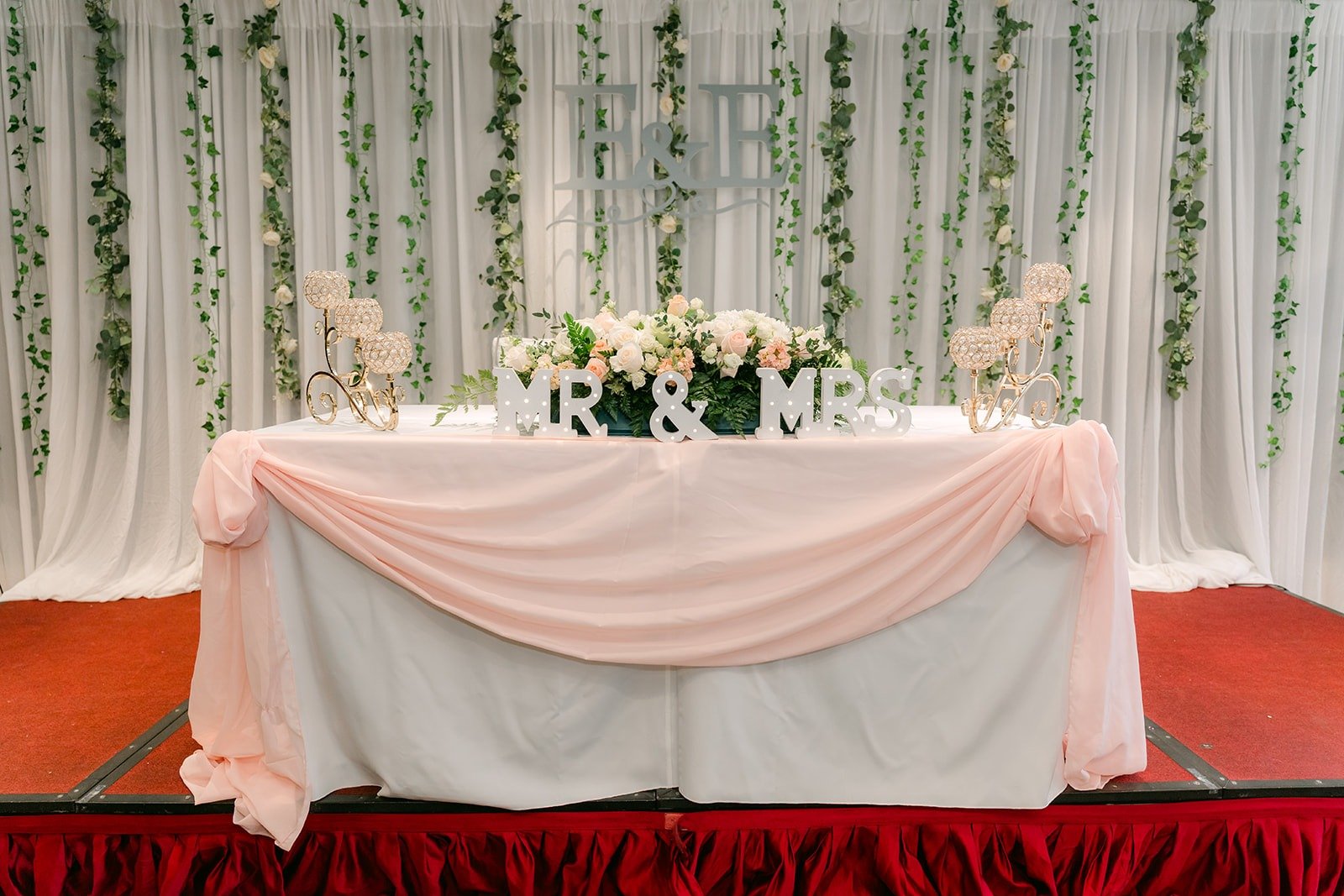 The sweetheart table draped in blush pink in front of a wall of greenery