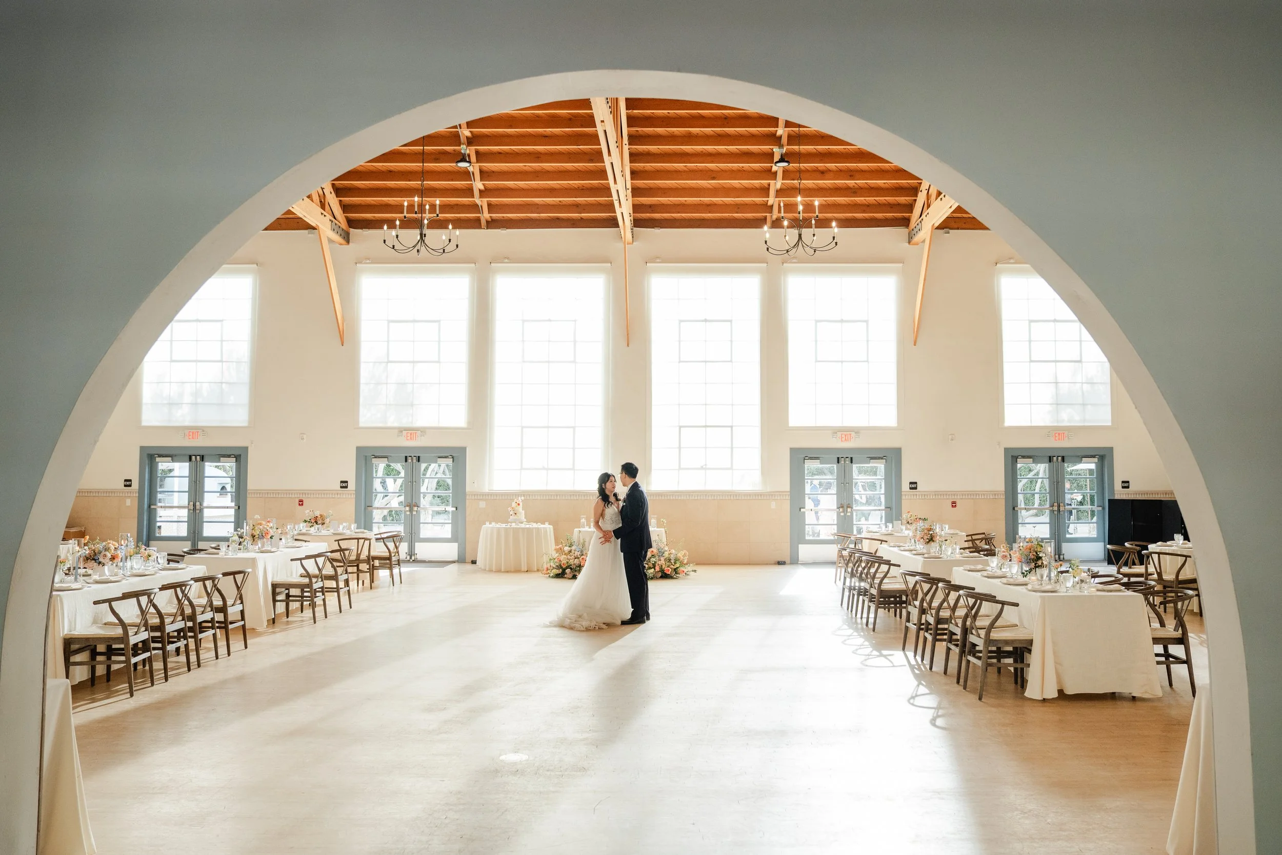 A bride and groom standing together in a decorated event hall with high windows and a wooden ceiling, wedding tables and chairs on either side, and floral arrangements, with sunlight streaming in.