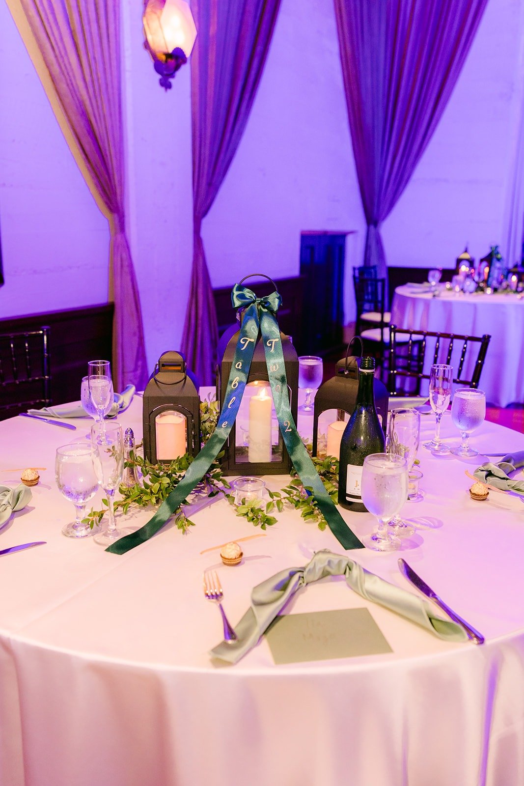 A round guest table set with sage napkins, lanterns and greenery