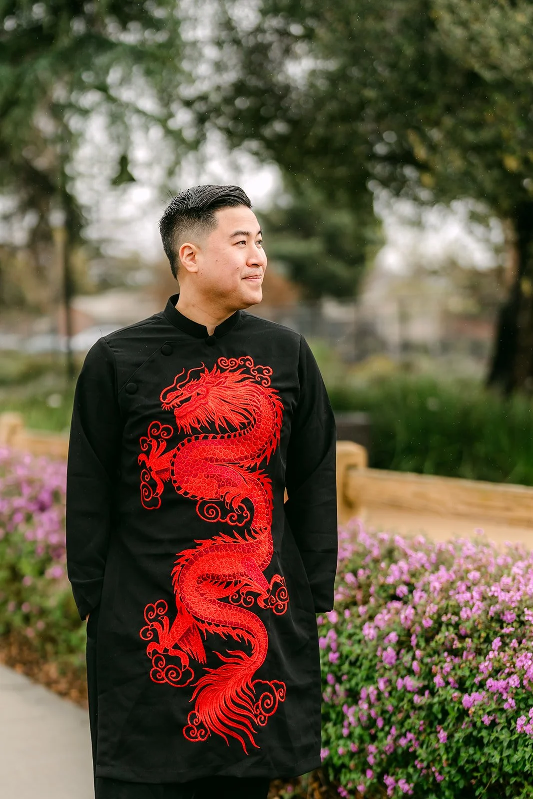 A groom in a black changshan with a red dragon embroidered down the front, standing in a garden.