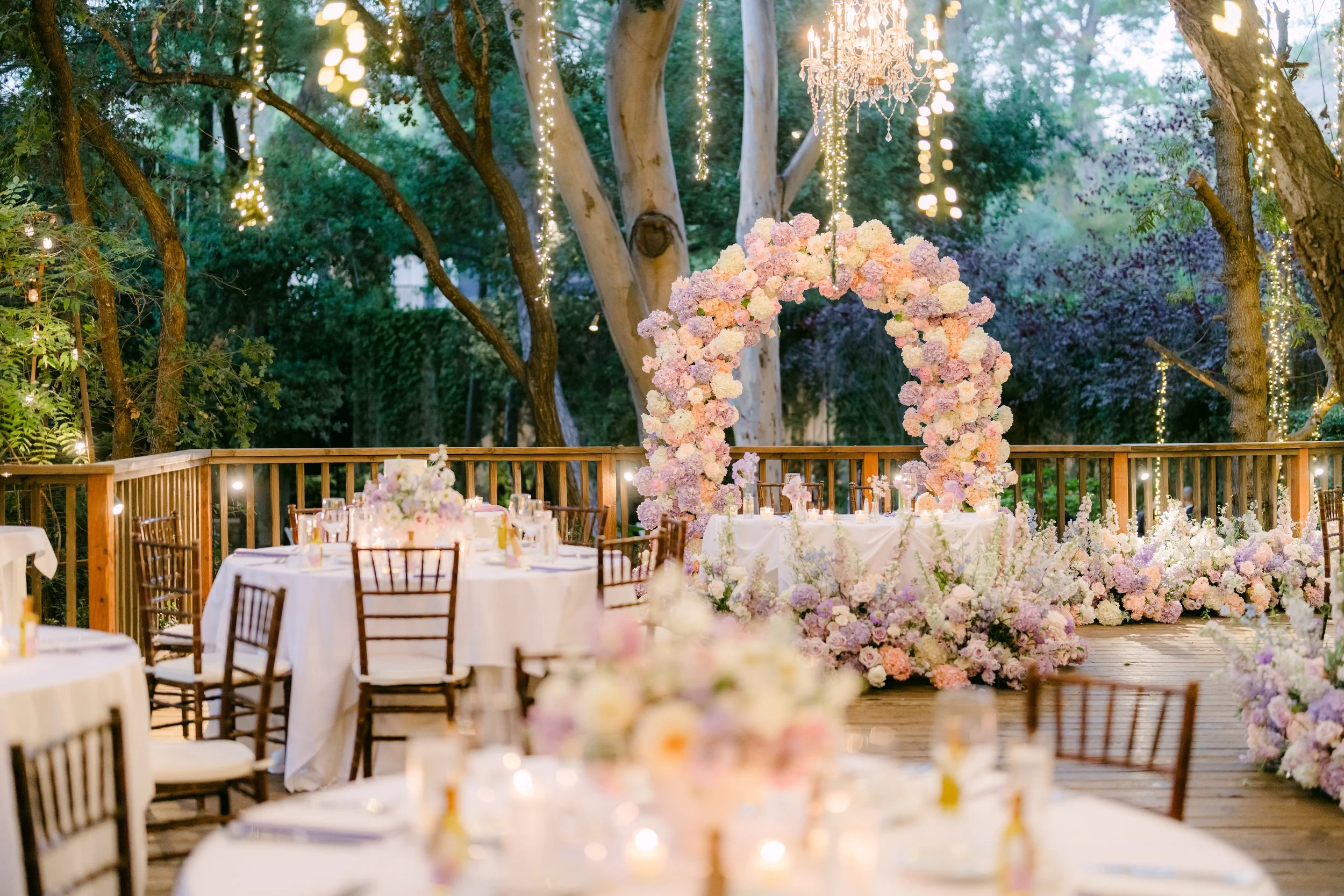 Outdoor wedding reception decorated with a large floral arch, string lights, and a chandelier overhead, with banquet tables and chairs set on a wooden deck surrounded by trees.