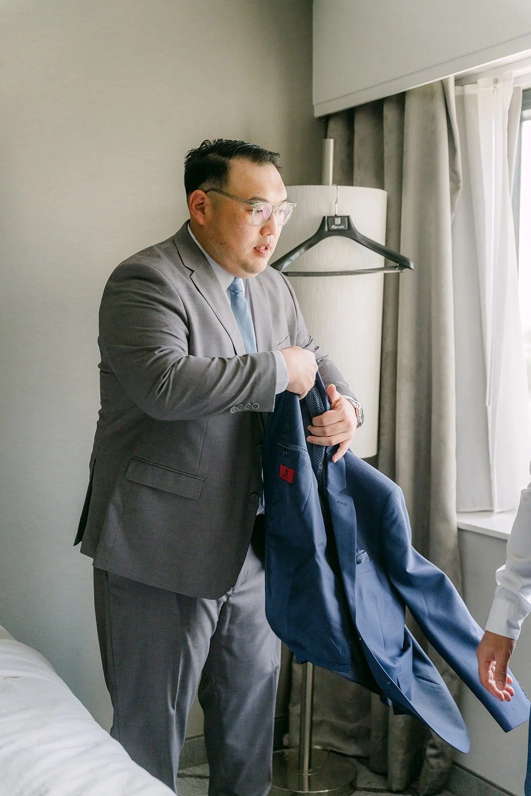 A groomsman holding up Mark's blue suit jacket in the hotel room