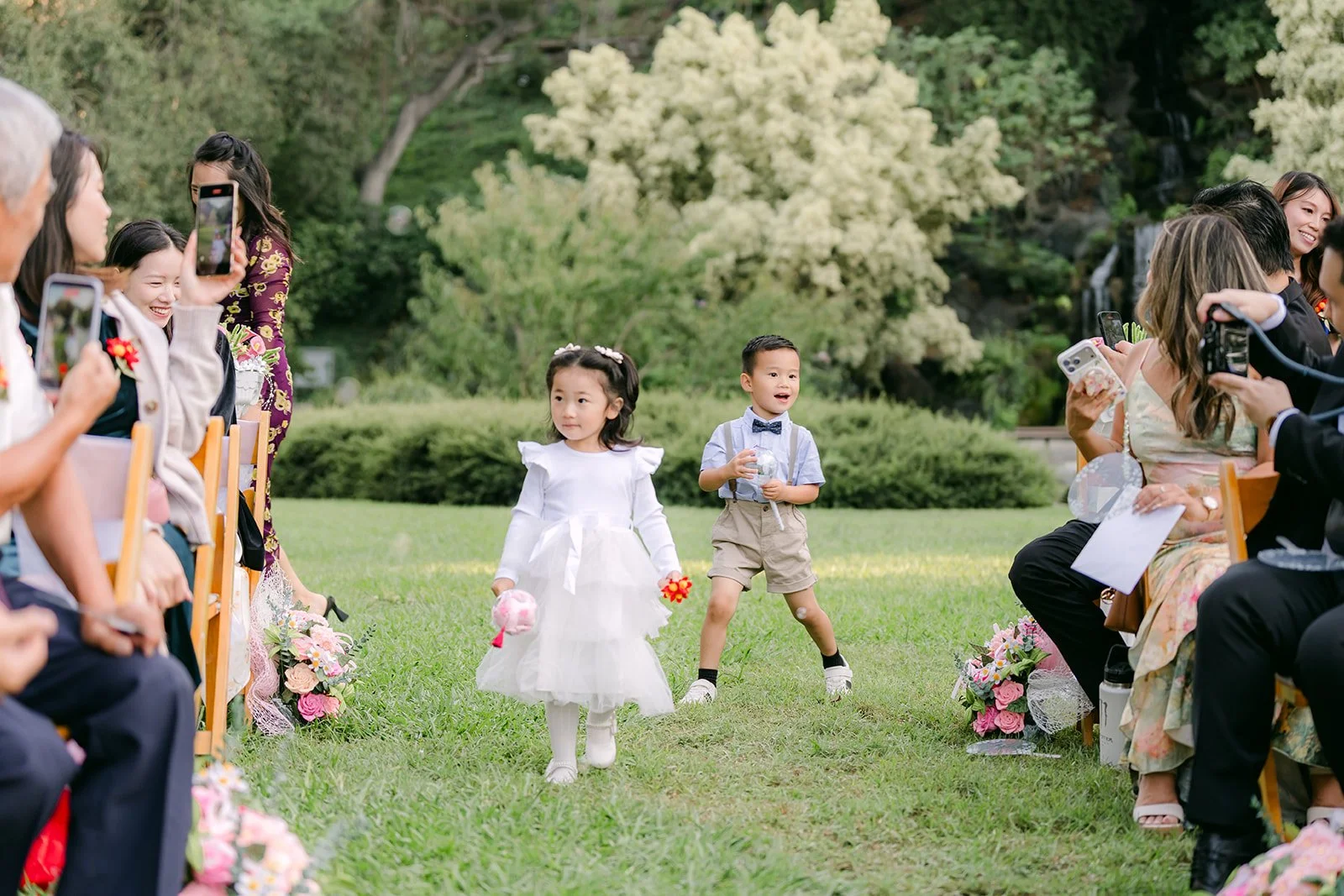 The flower girl and ring bearer walking down the aisle as guests film.