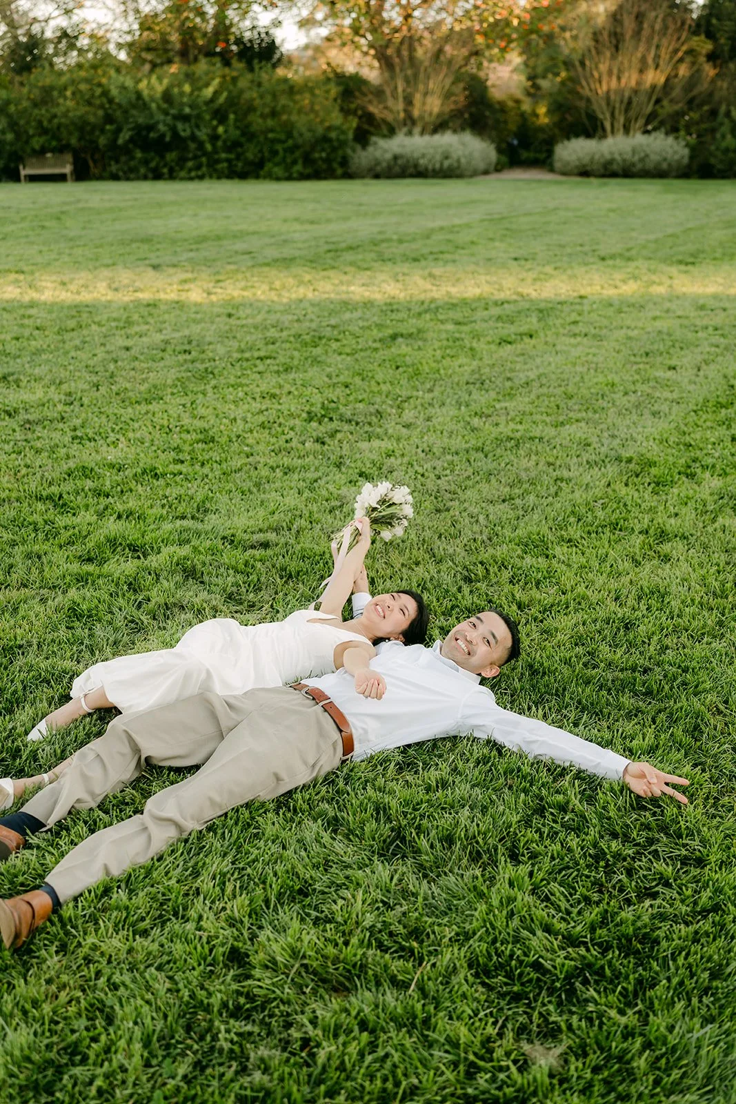 Kim and Vincent lying on the grass holding up her bouquet.