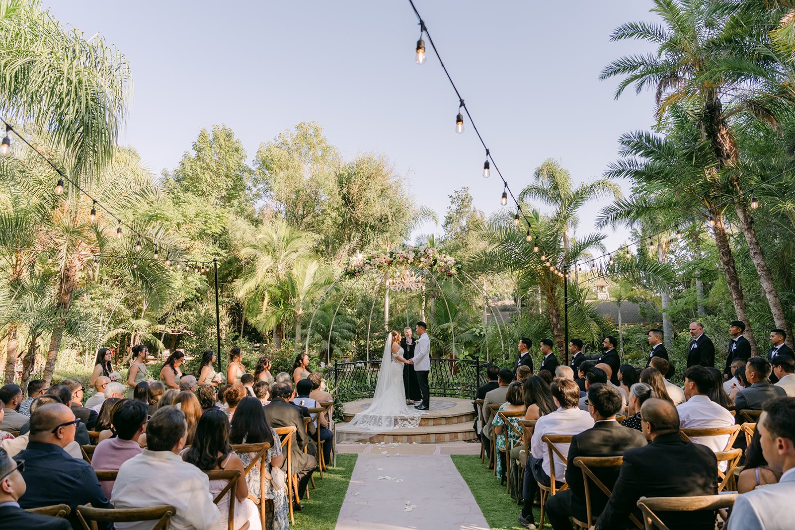 Wide view of Nicole and Caryl's ceremony under the string lights at Eden Gardens.