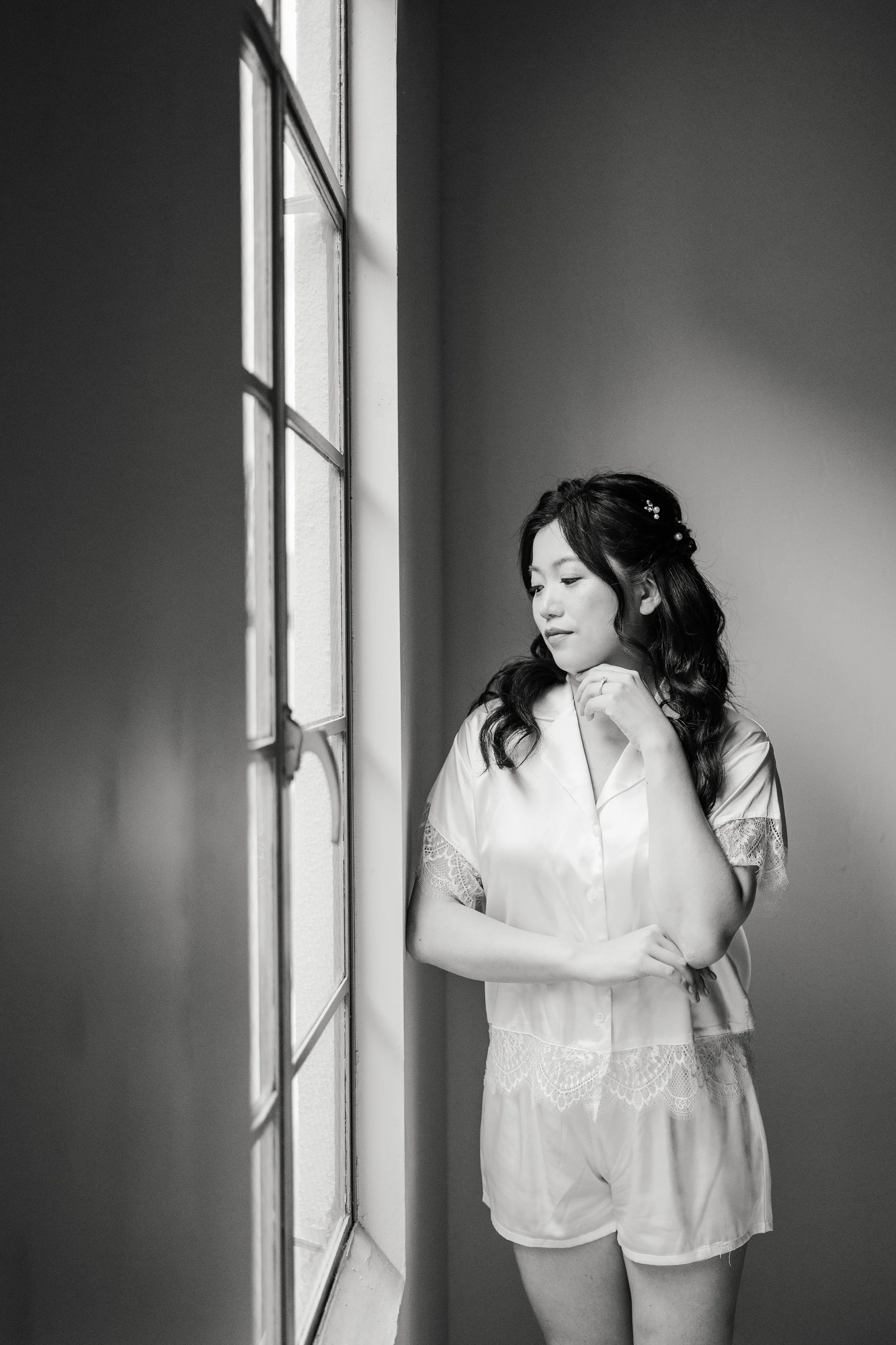 A woman with long, dark hair styled with decorative pearl hairpins is looking out a window. She is standing with her arms crossed near her waist, wearing a satin short-sleeved blouse with lace trim and matching shorts, captured in black and white.