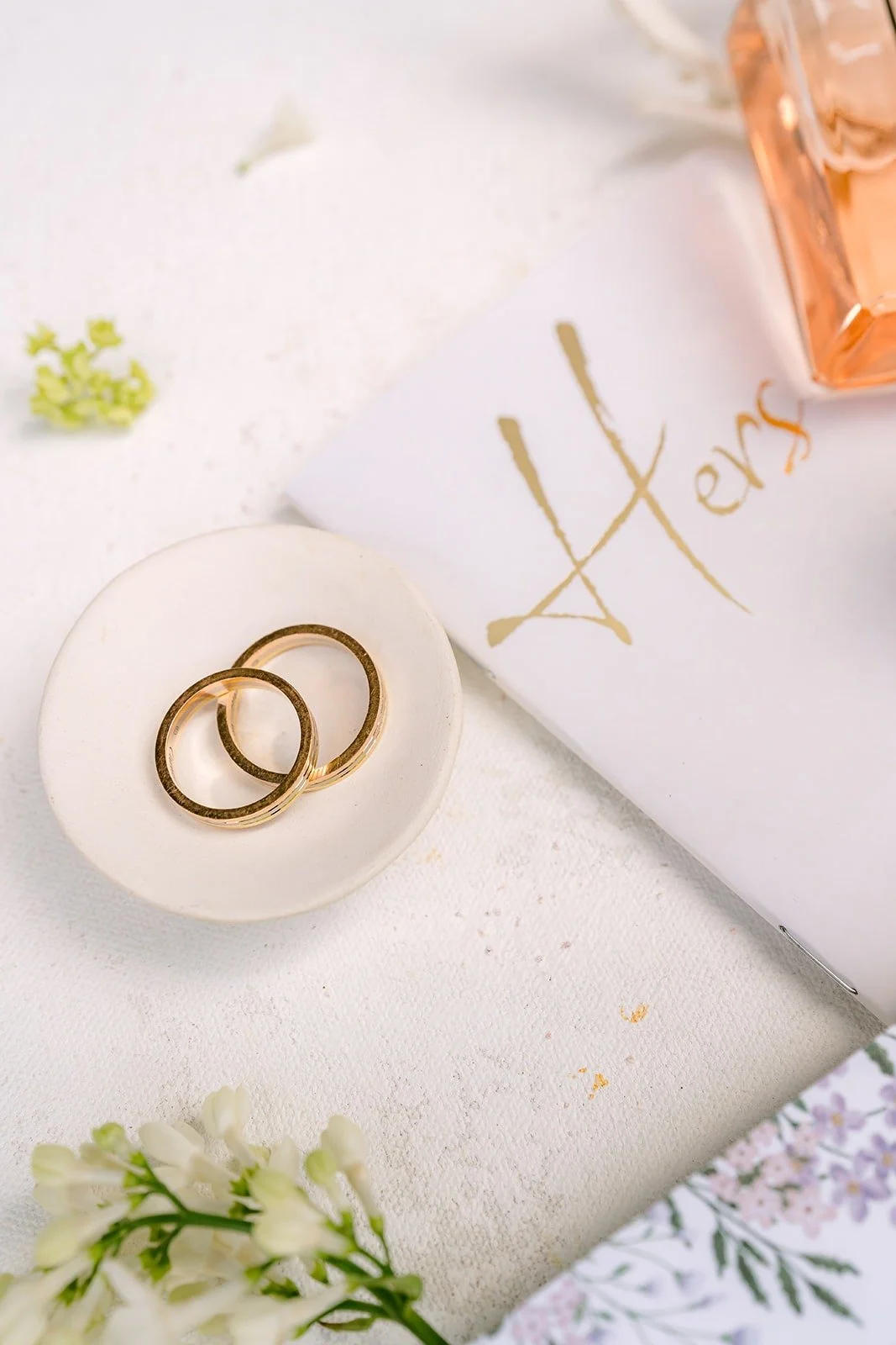 Two wedding bands resting on a card lettered "His" beside white flowers