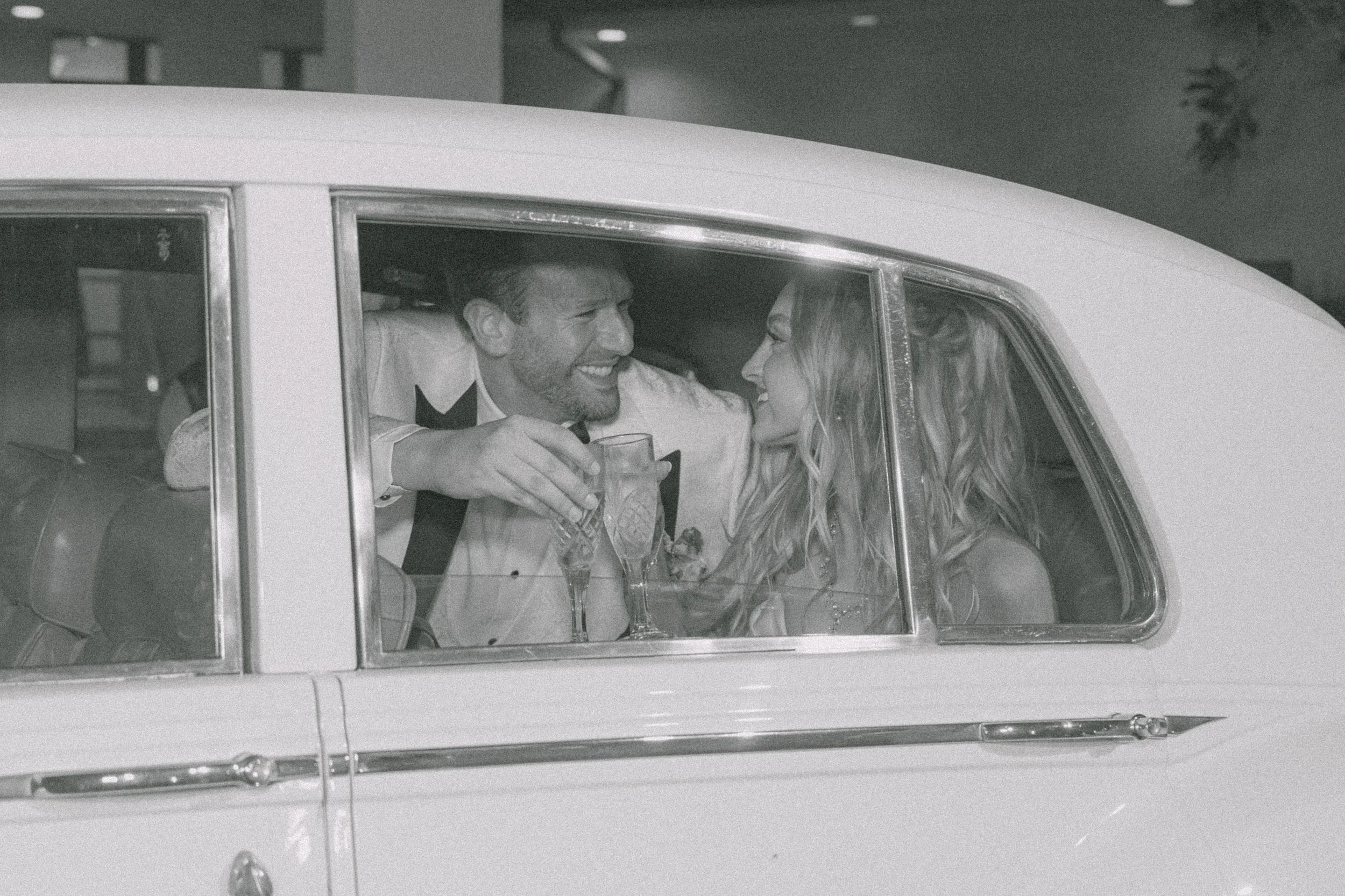 Black and white photo of a man and woman smiling and leaning close to each other in a car, holding glasses, during nighttime.