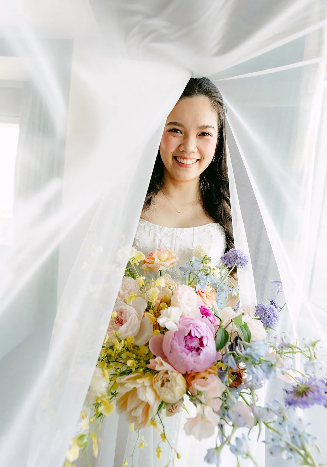 Linh smiling through her veil with a pastel peony bouquet