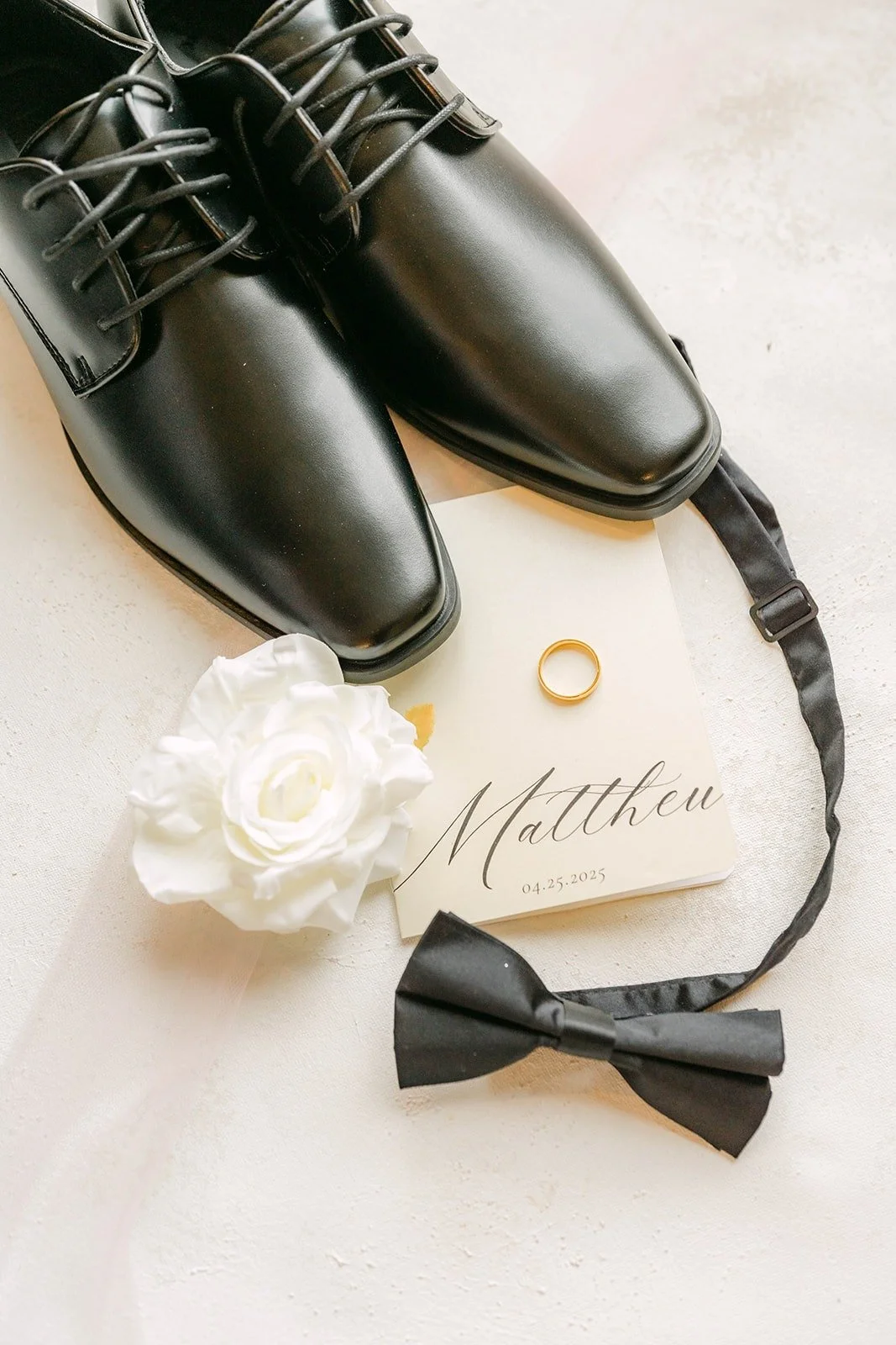 Matthew's black dress shoes, bow tie, wedding band and a white rose on a card with his name