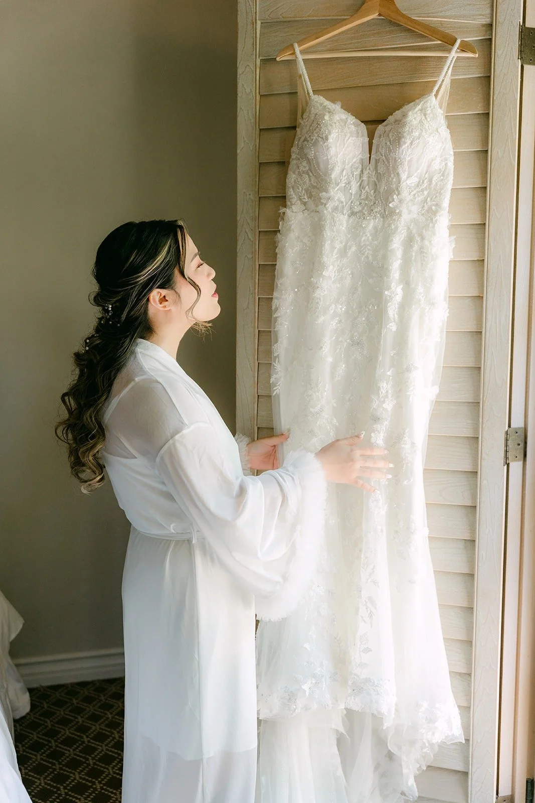 Joanne in a white robe looking at her wedding gown hanging by the window