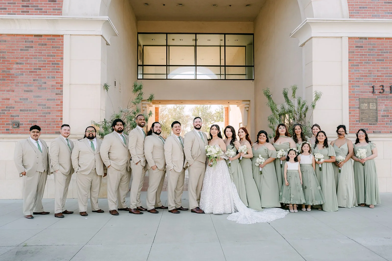 The full wedding party in sage and tan in front of a brick building.