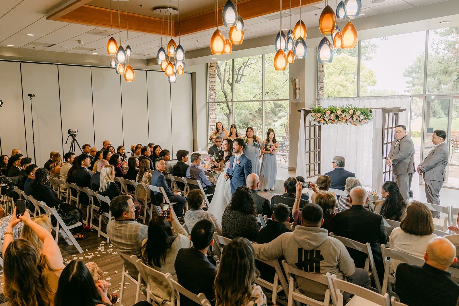 The ceremony room at Rio Hondo filled with guests and views of the trees through the windows, photographed by One and Only Production