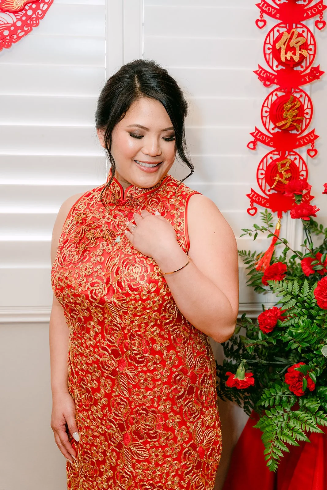 A bride in a red and gold qipao standing beside double happiness couplets.