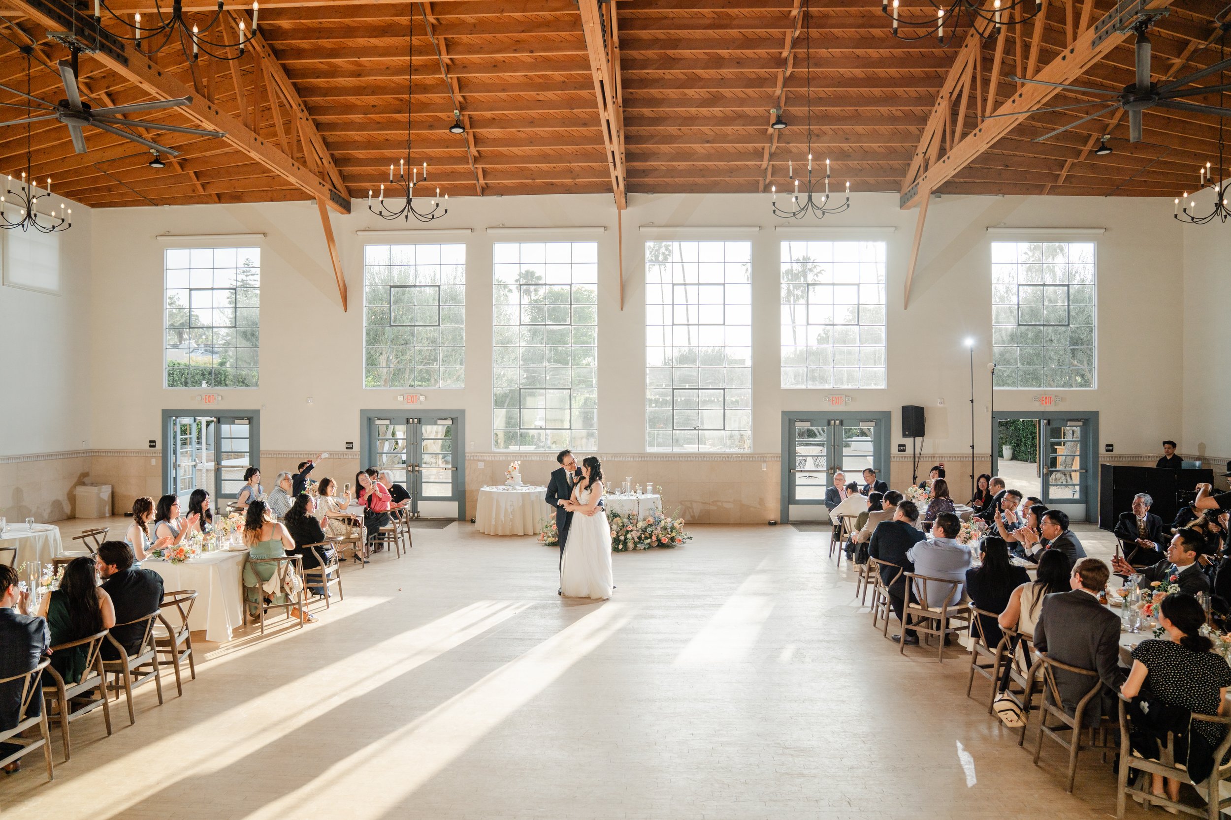 A wedding reception in a spacious hall with high wooden ceiling, large windows, and chandelier lighting. The bride and groom are standing in the center, facing each other, surrounded by seated guests. The guests are seated at round tables decorated w