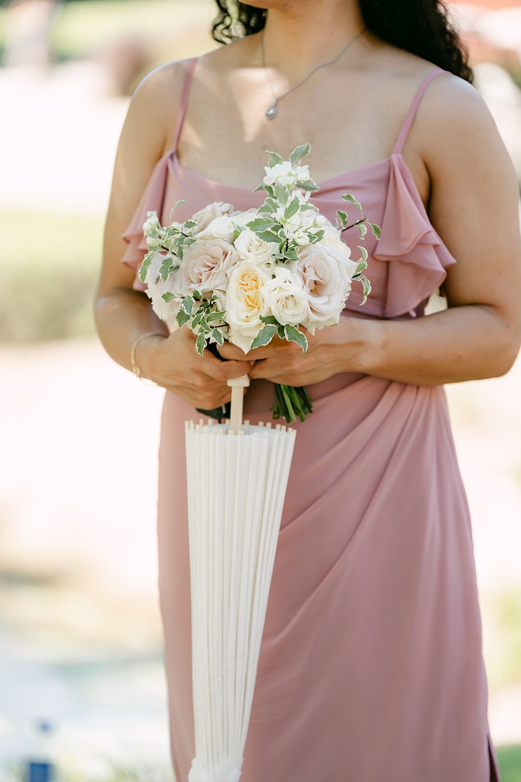 A bridesmaid in a dusty pink gown holding a white umbrella and bouquet.