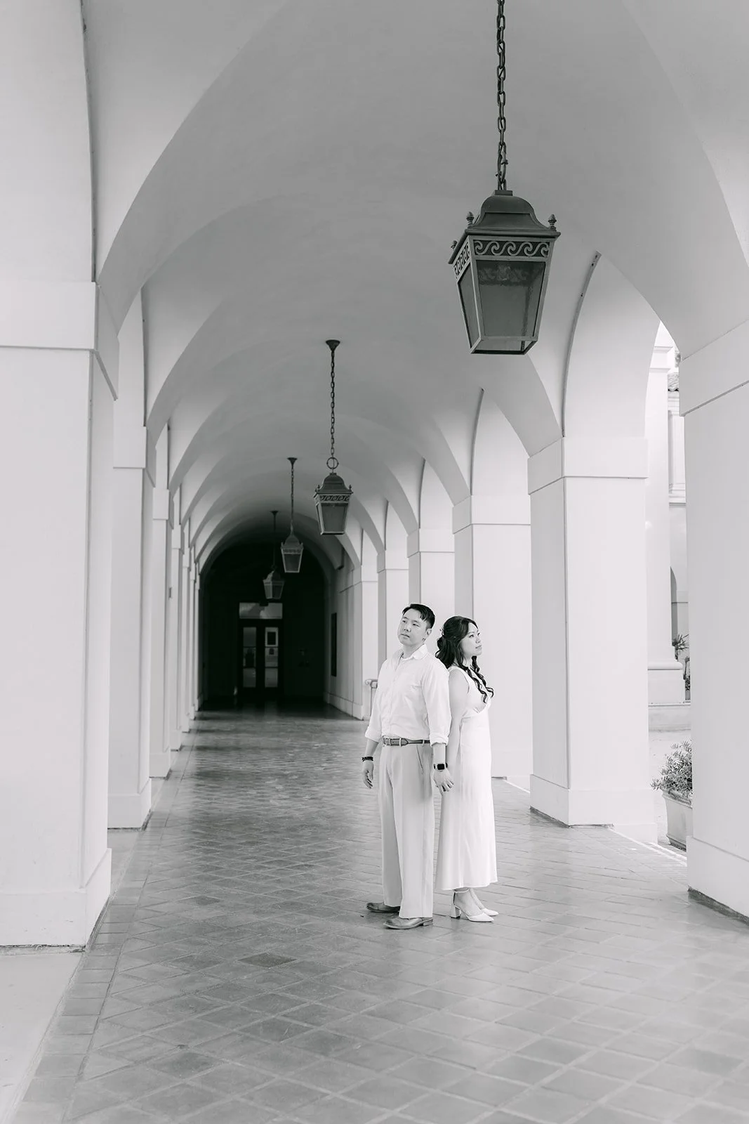 Black and white photo of Emily and James standing in the long arched colonnade at Pasadena City Hall.