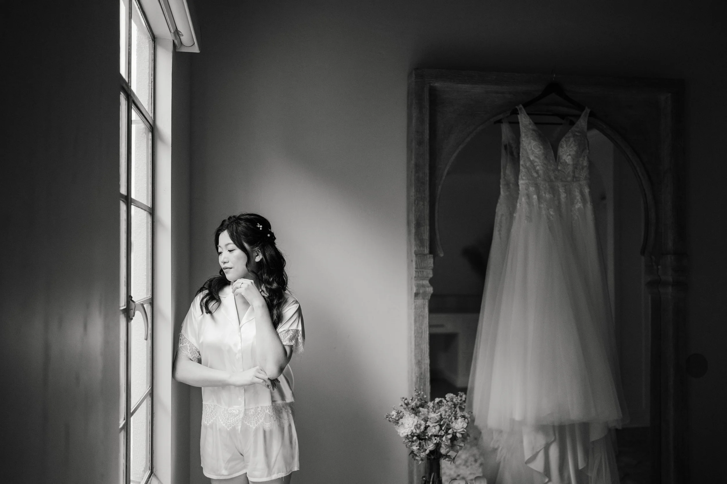 A woman with long dark hair, wearing a light-colored robe with lace sleeves, standing near a window in a room with a wedding dress hanging on the wall. She appears to be deep in thought or prayer. There is a bouquet of flowers on a table nearby.