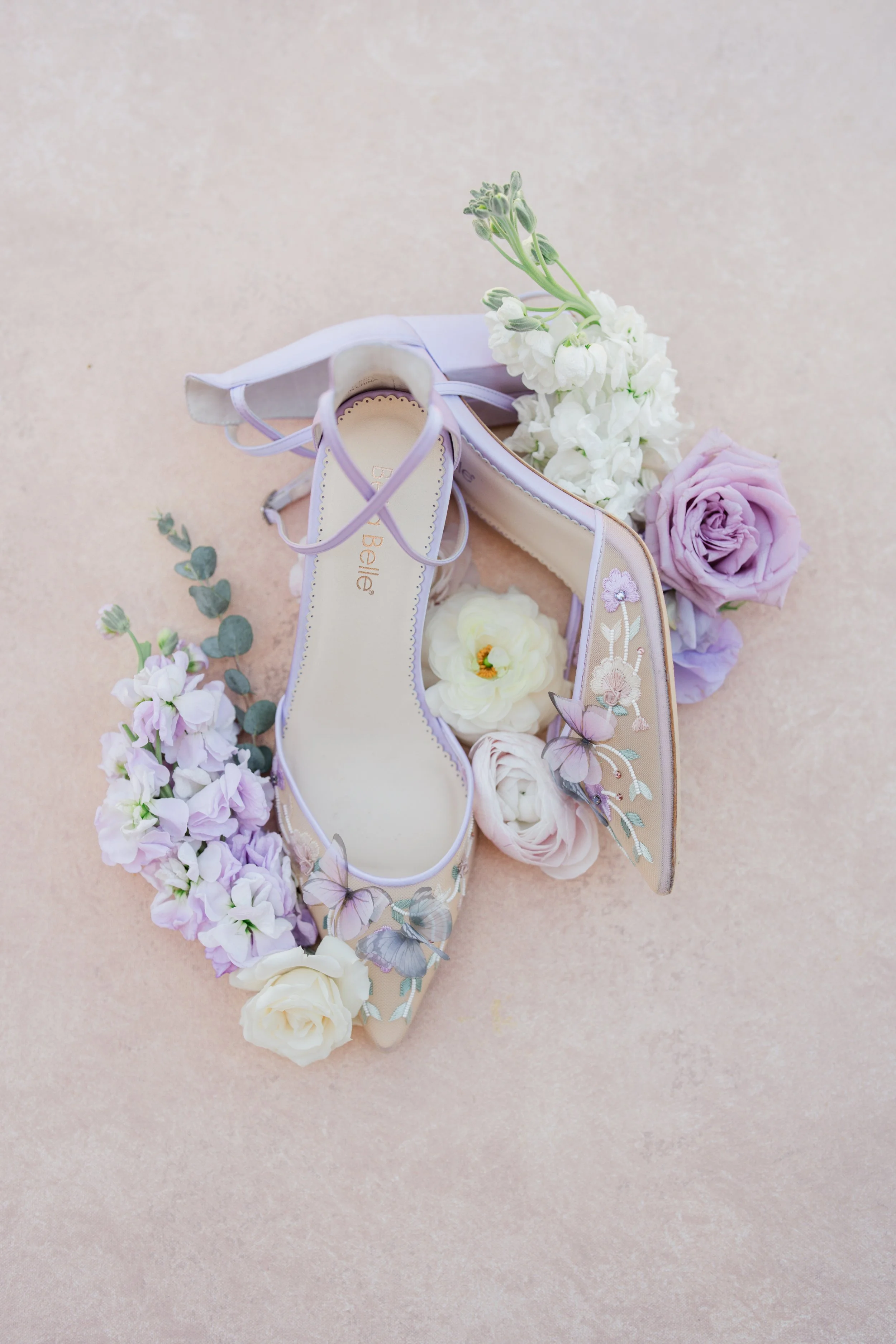 A pair of light purple floral embroidered high-heeled shoes surrounded by white, pink, and lavender flowers on a soft pink background.