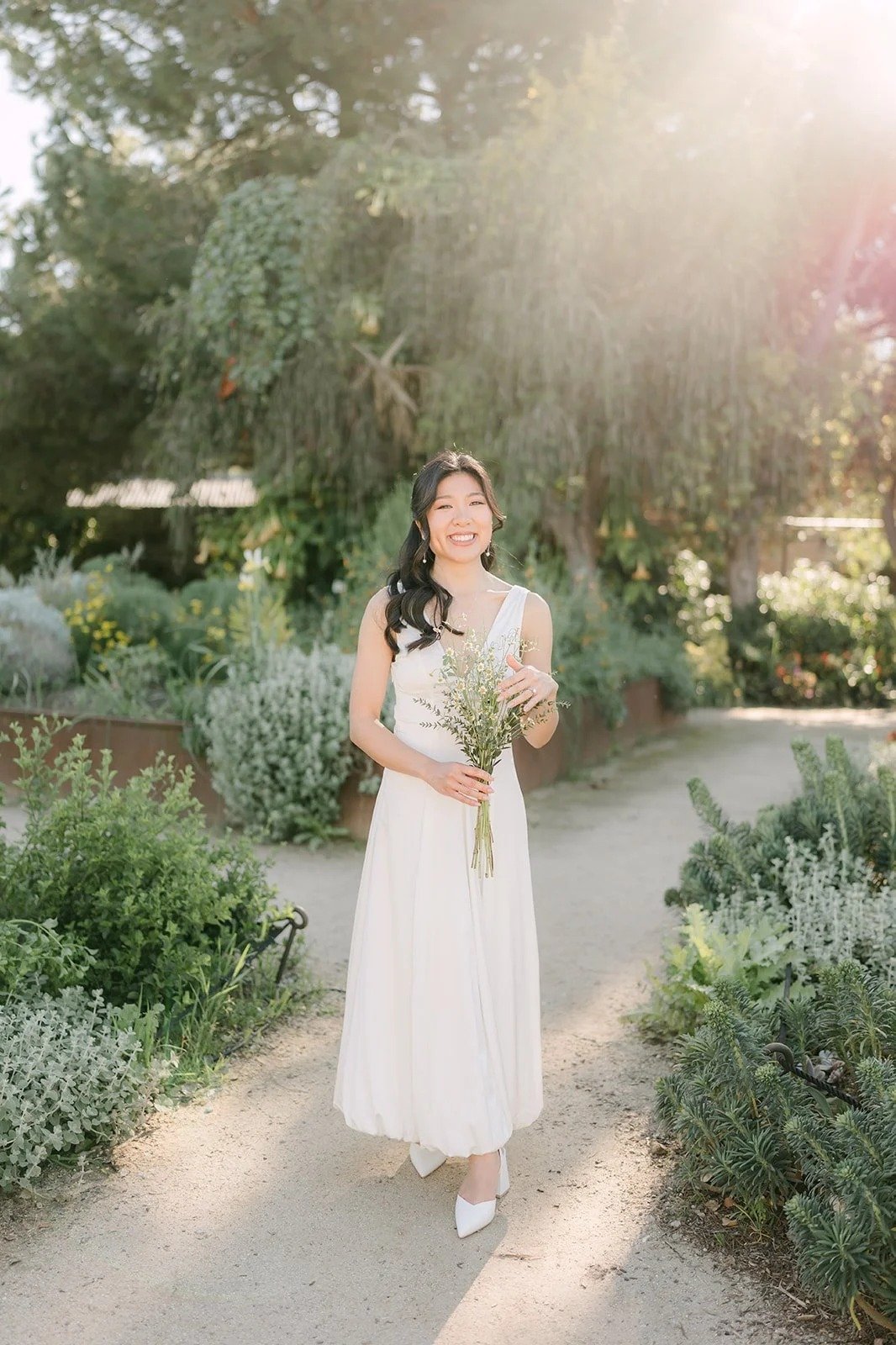 Kim in a white V-neck dress holding a wildflower bouquet on a sunlit garden path at South Coast Botanic Garden.