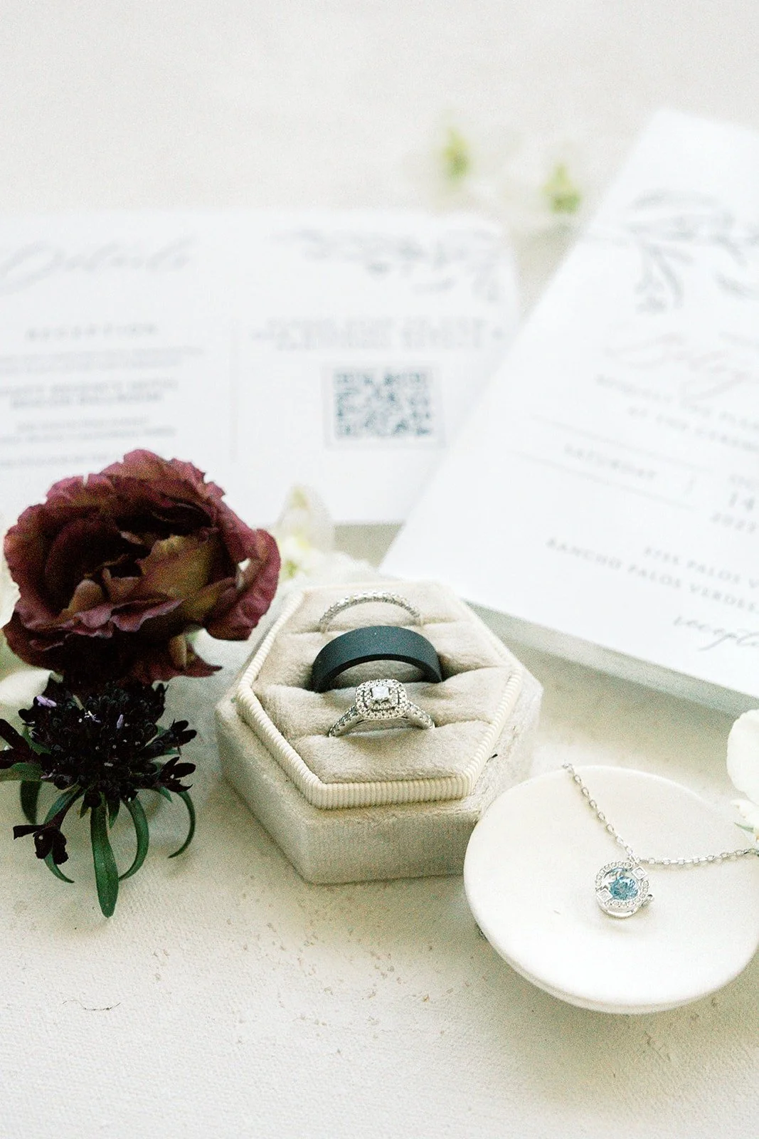 Wedding rings in a cream ring box beside a pendant necklace and burgundy ranunculus