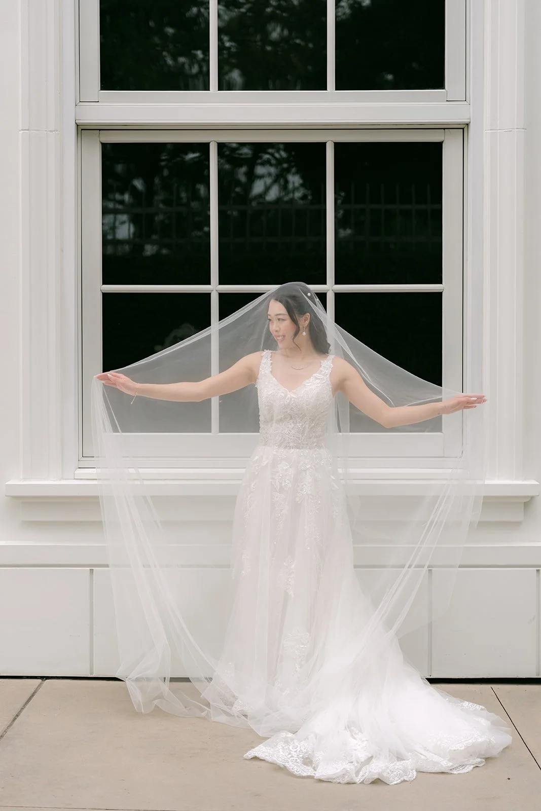 Jenny spreading her veil in front of a white wall and window.