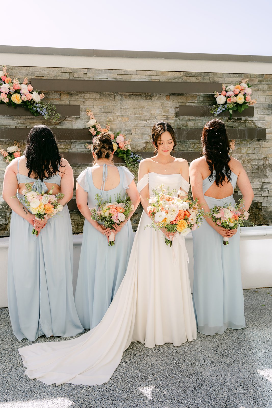 Amanda and her bridesmaids lifting their bouquets in front of the stone wall.