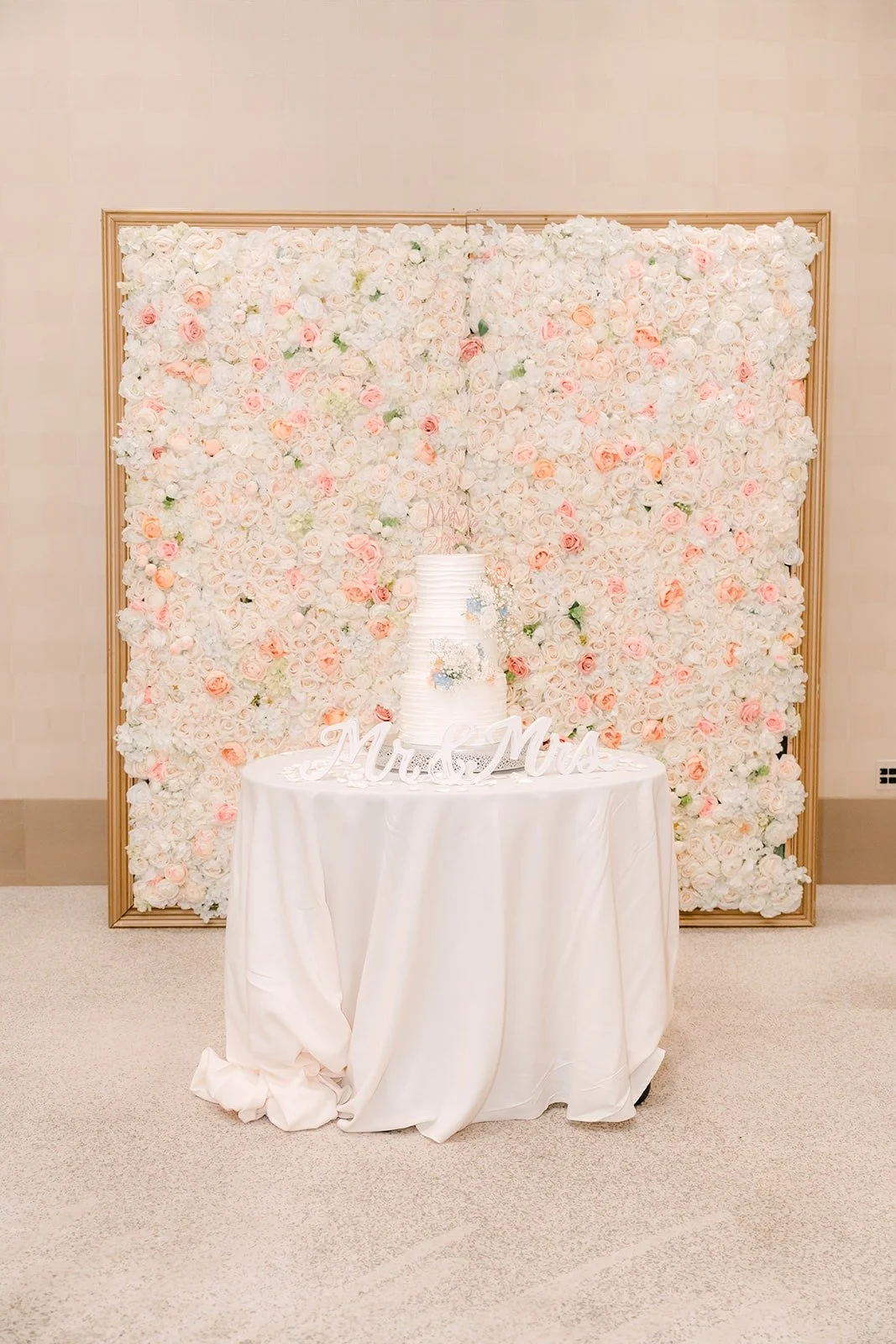 The white wedding cake in front of a flower wall in the Carousel Ballroom