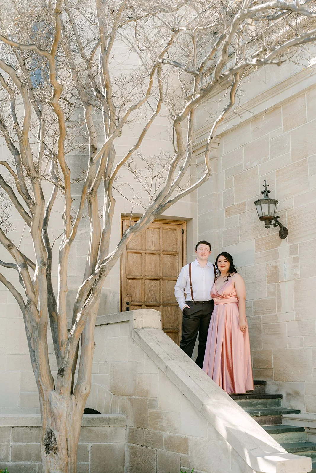 The couple at the mansion's wooden door beneath a sculptural bare tree.