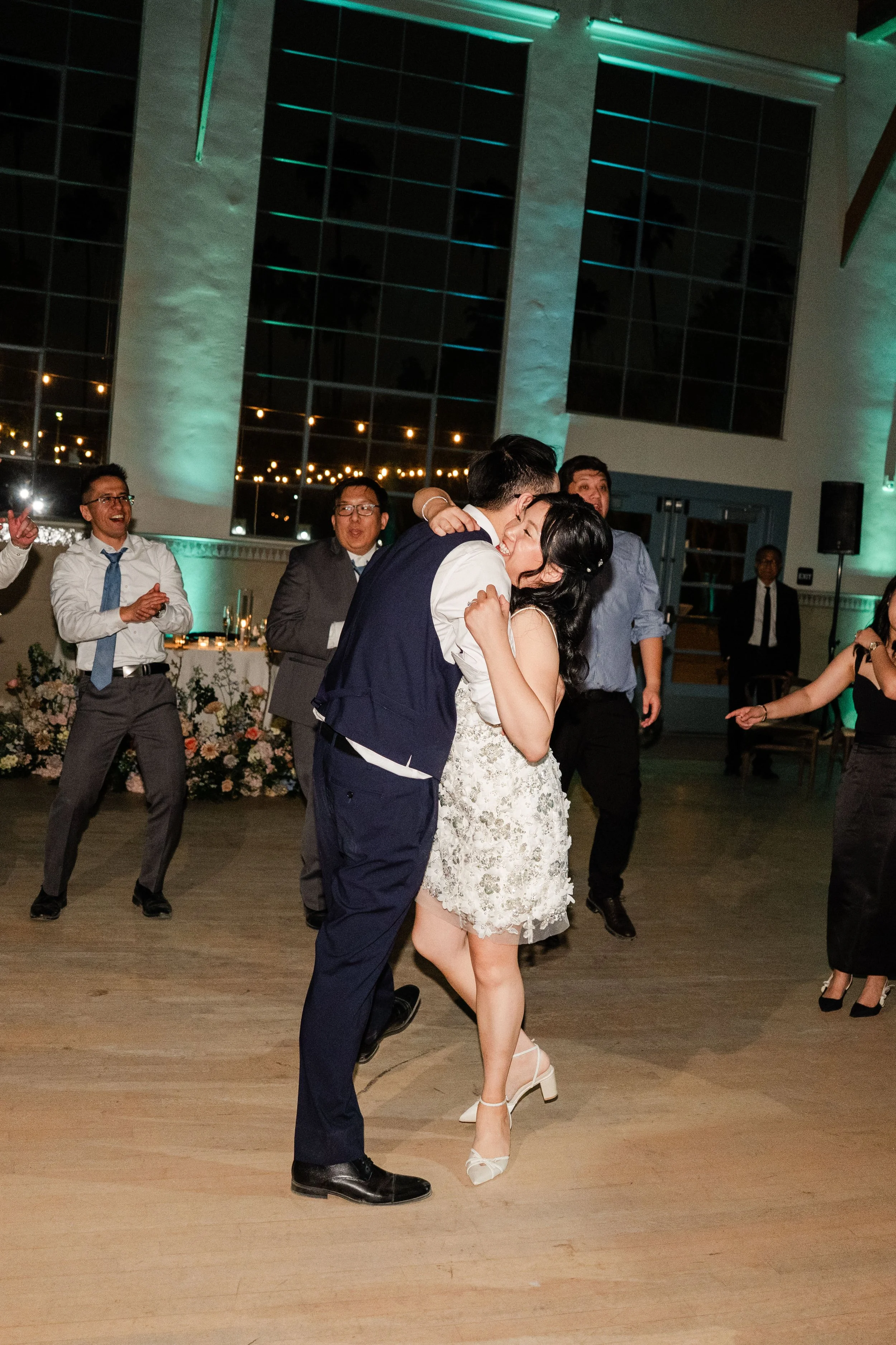 A group of people dancing and celebrating at a wedding reception, with two people sharing a dance in the center, as others watch and cheer around them, in a decorated indoor venue with large windows and string lights.