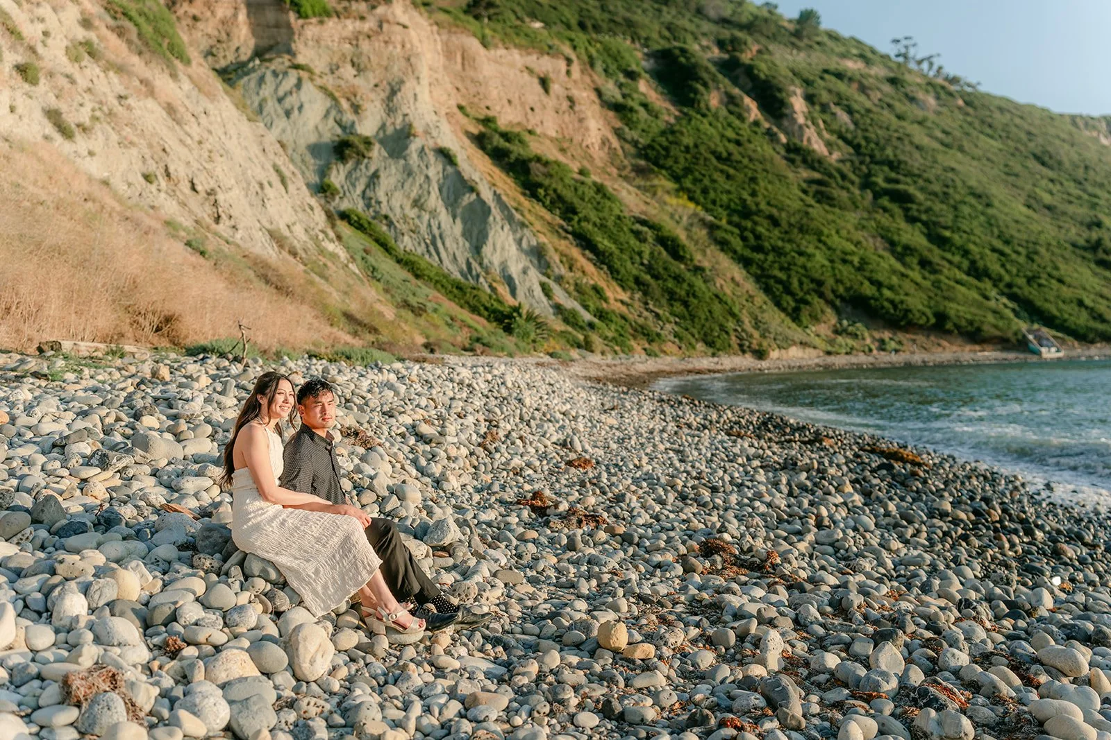 Victoria and Andrew sit together on the rocky shore below the green hillside, photographed by One and Only Production.