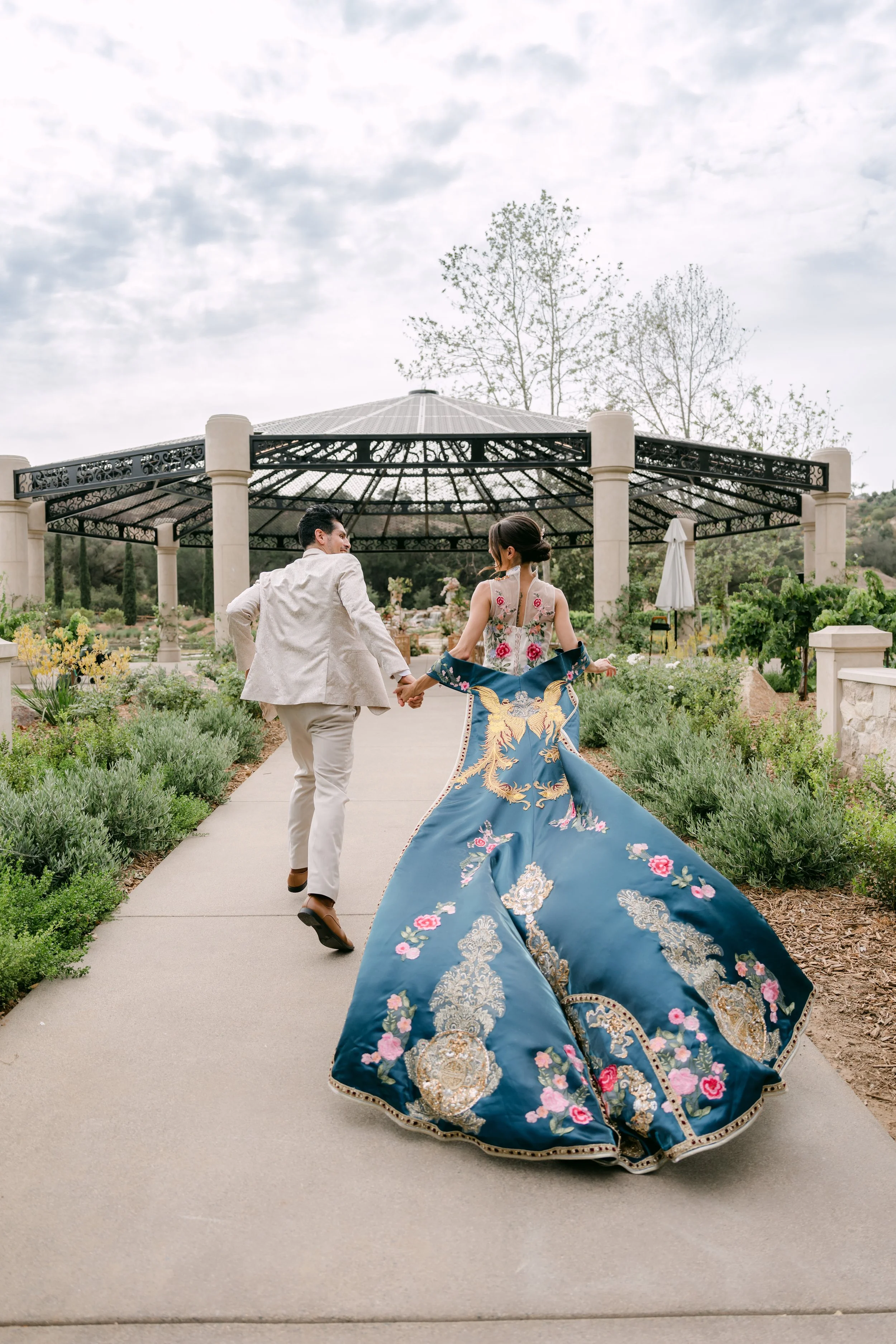 Couple running down a garden path, her blue embroidered gown trailing, at Monserate Winery