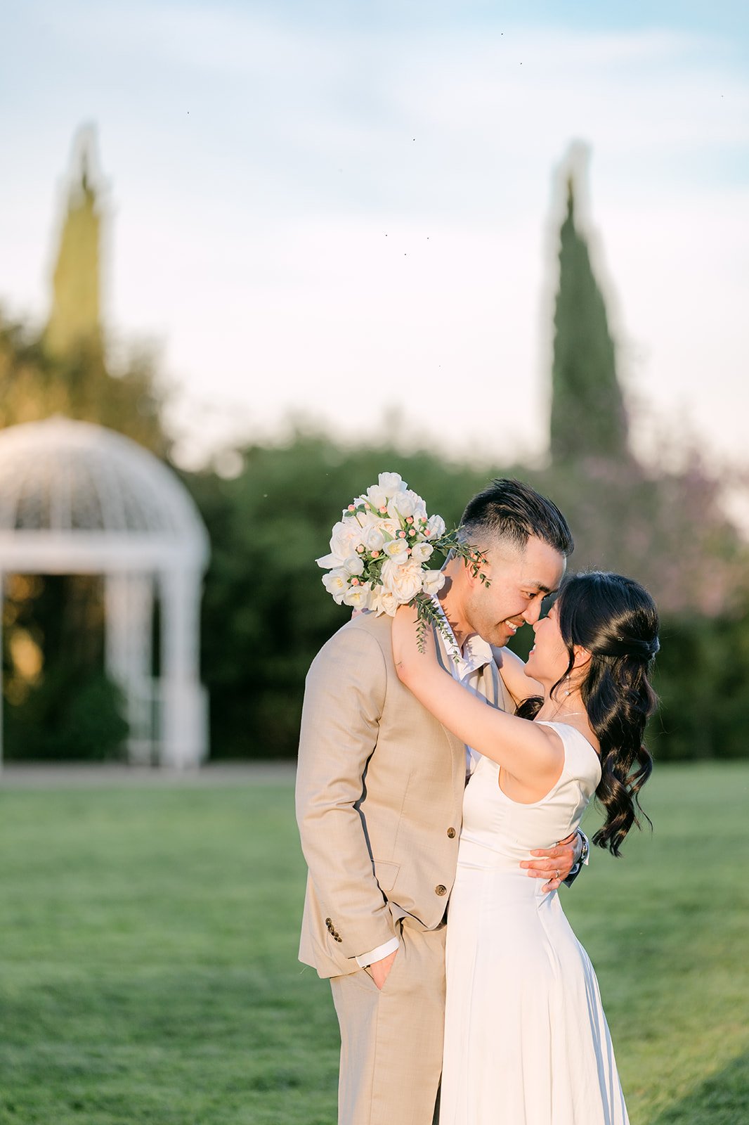 Kim and Vincent kissing behind her bouquet with the gazebo and cypress trees beyond, photographed by One and Only Production.