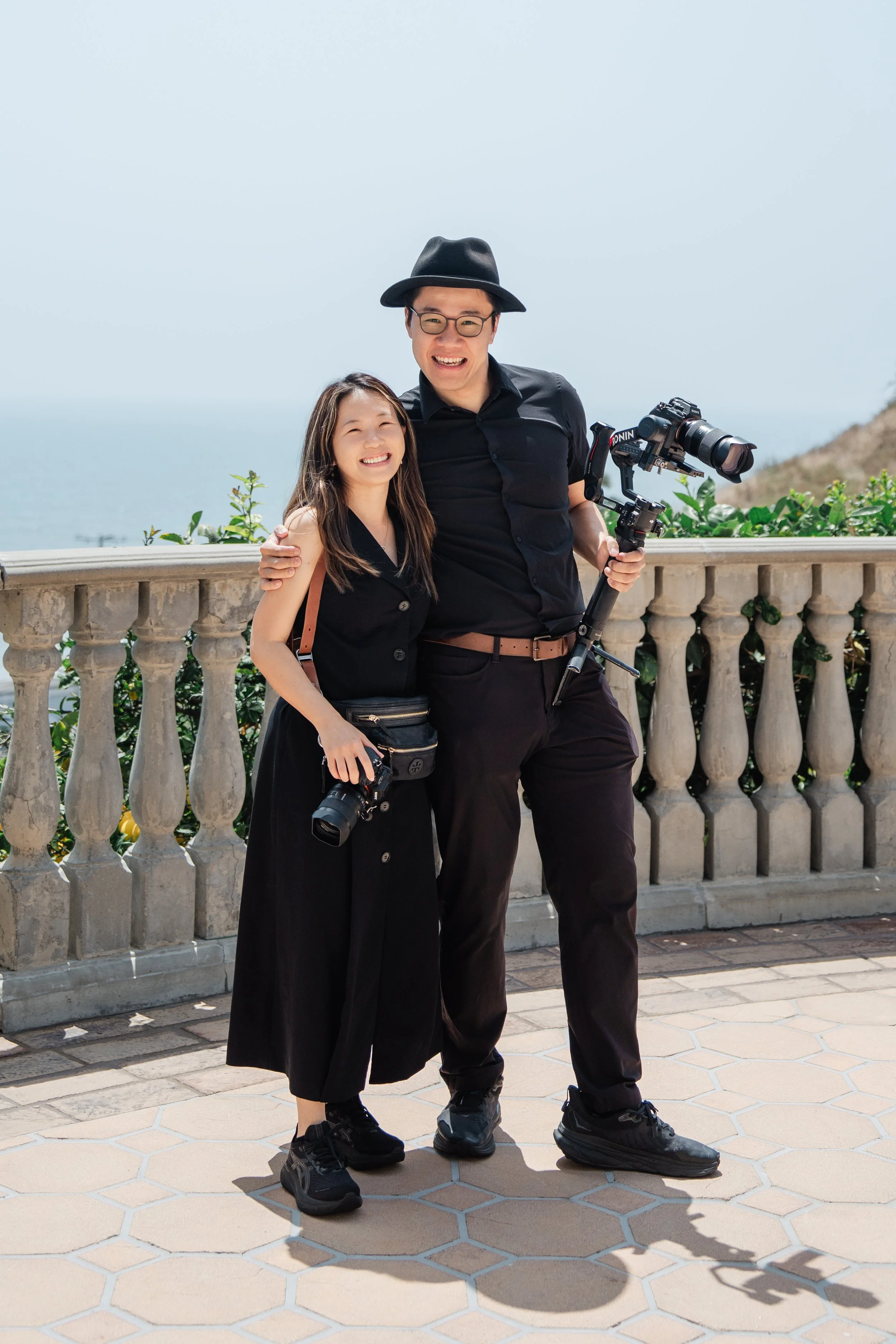 Two friends, a woman and a man, smile and pose together on a sunny outdoor balcony with a scenic ocean view in the background. They are dressed in black and holding cameras, with the man also holding a stabilizer for his camera.