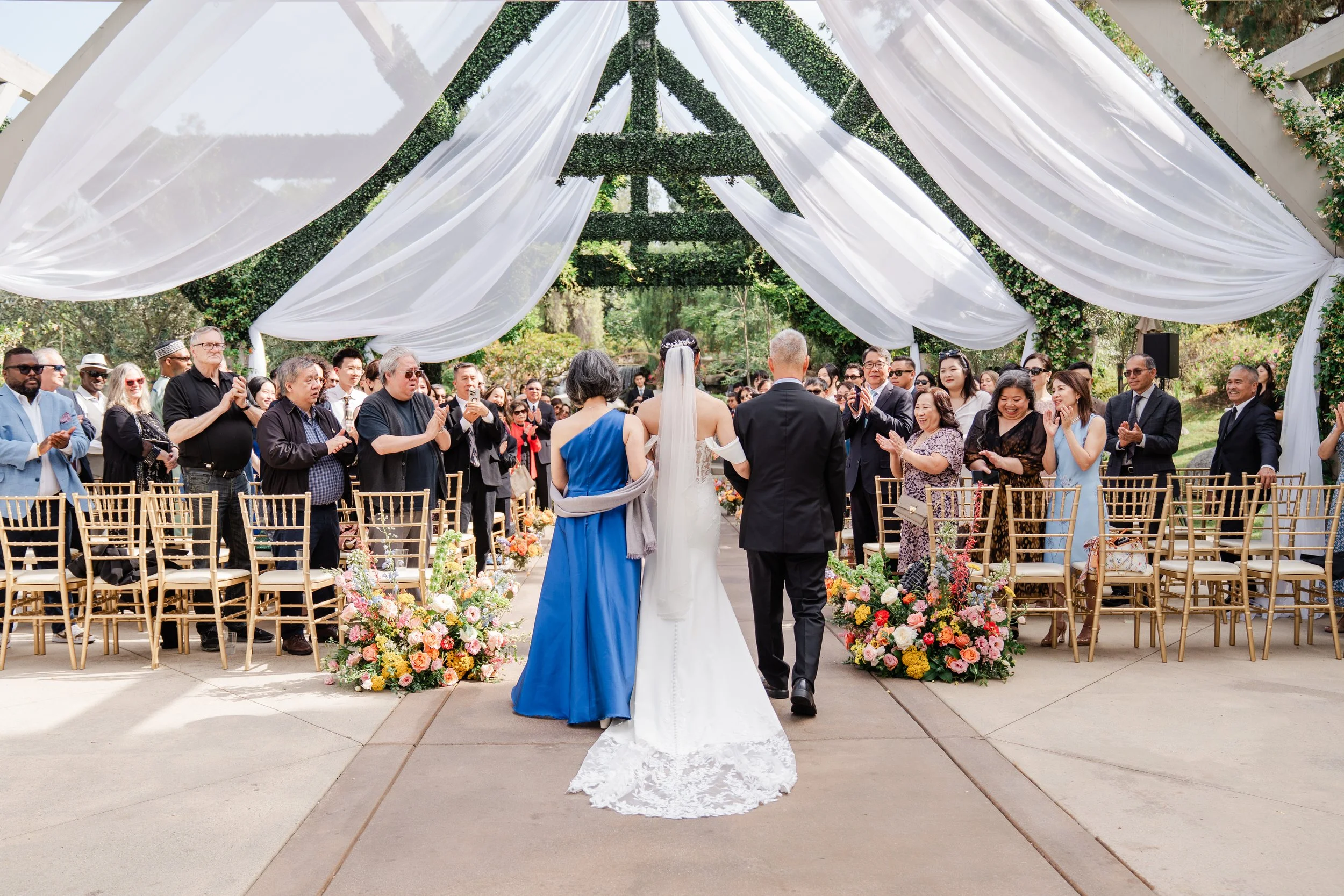 A wedding ceremony outdoors with a bride and groom standing under a canopy decorated with white drapery and greenery, surrounded by seated guests.