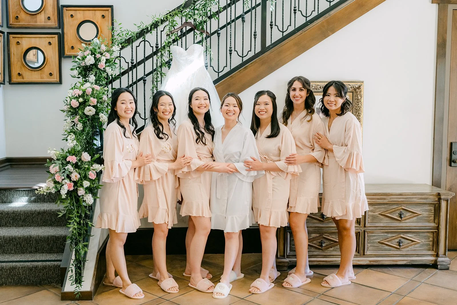 Bride and bridesmaids in matching blush robes posing by the staircase at Vellano Estate