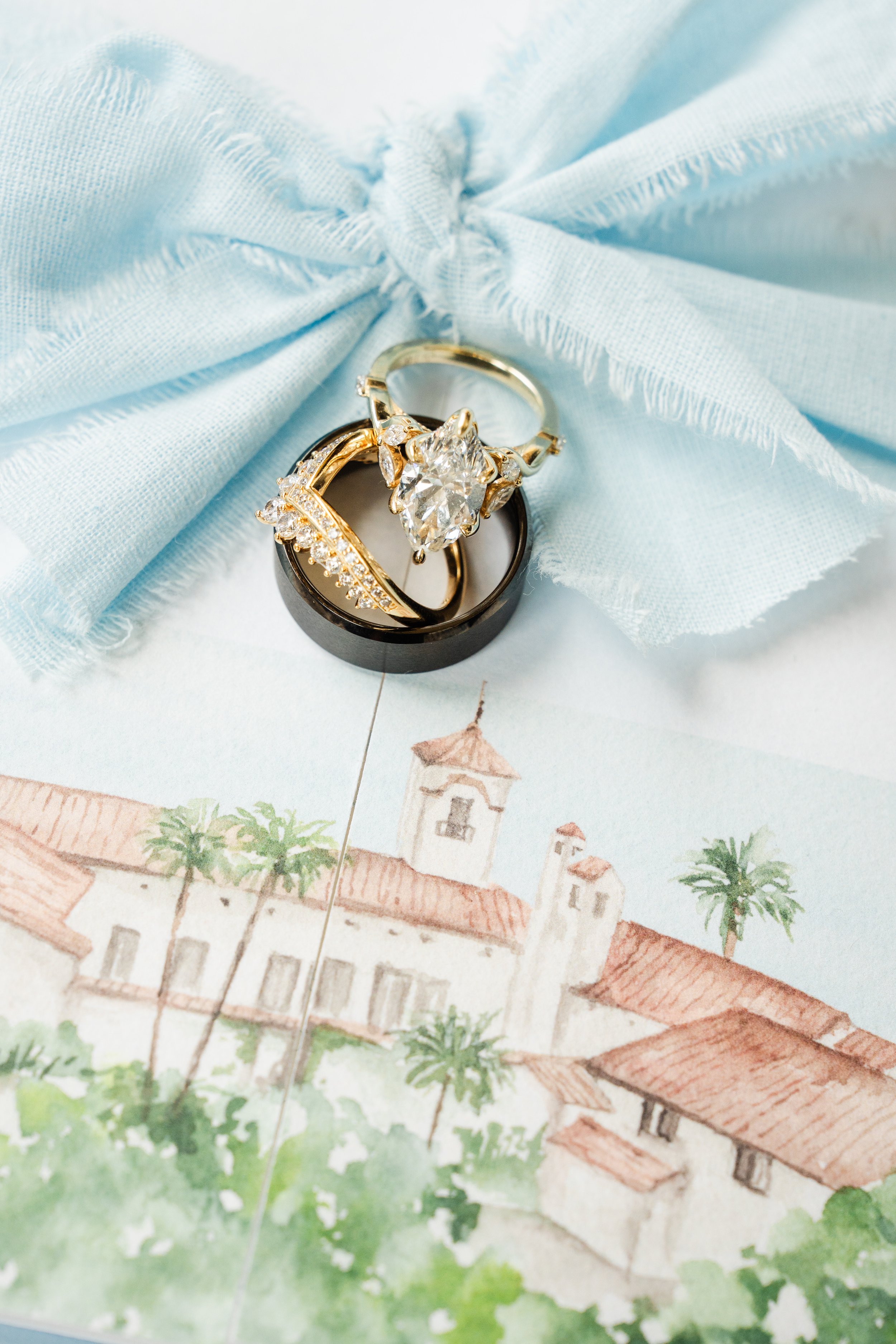 Close-up image of two rings resting on a black circular box, all placed on a light blue fabric with frayed edges. One ring is gold with a large central clear gemstone, and the other is gold with smaller clear gemstones.