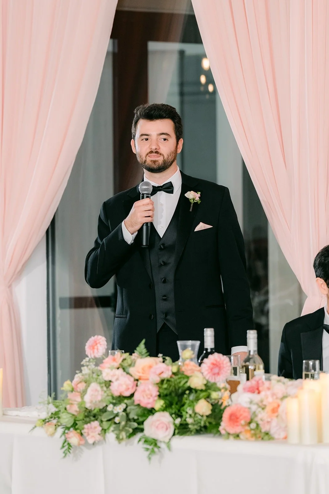 A guest in a tuxedo giving a toast in front of the blush drapes