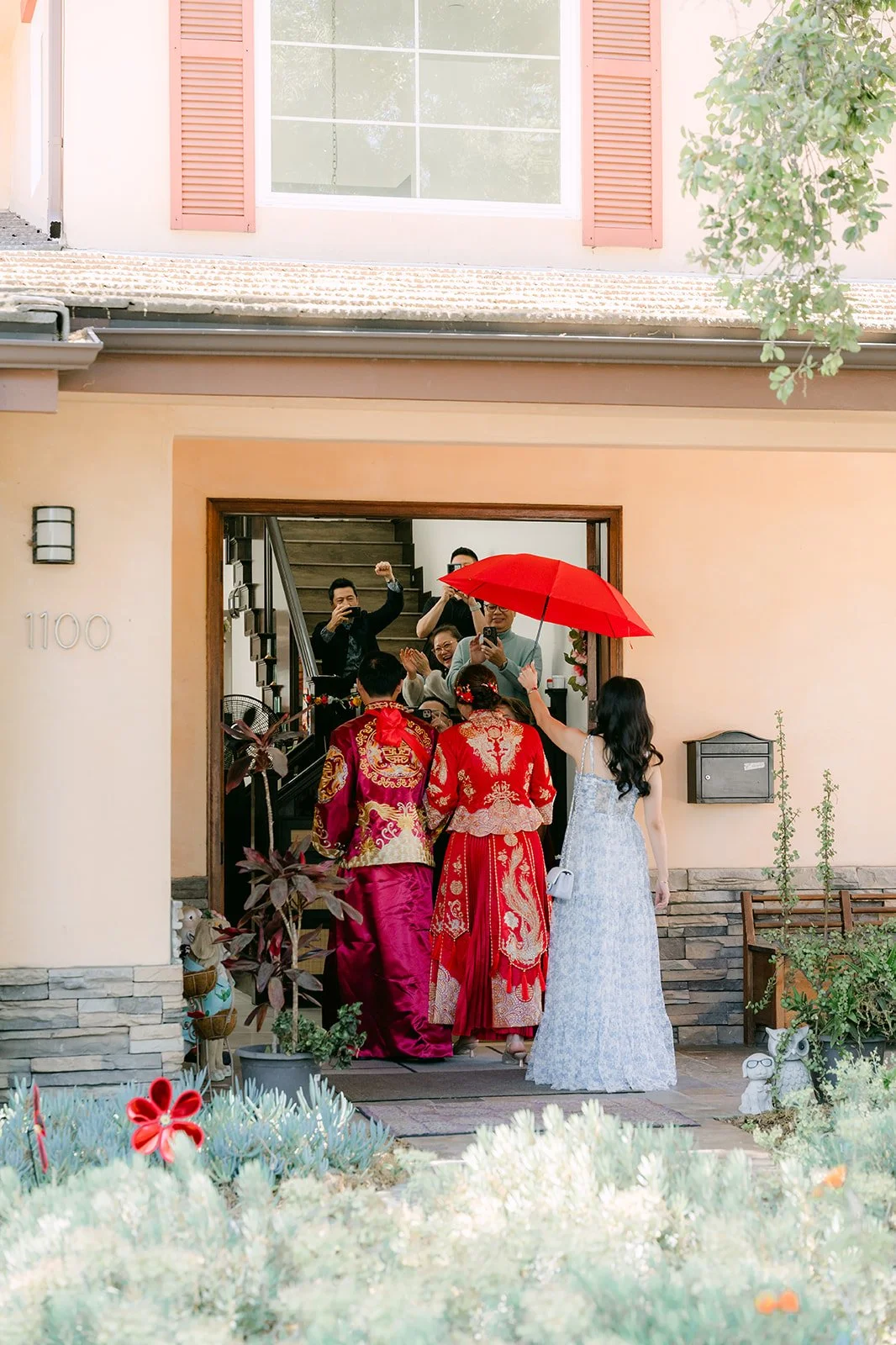 The couple leaving the front door under a red umbrella with family behind them.