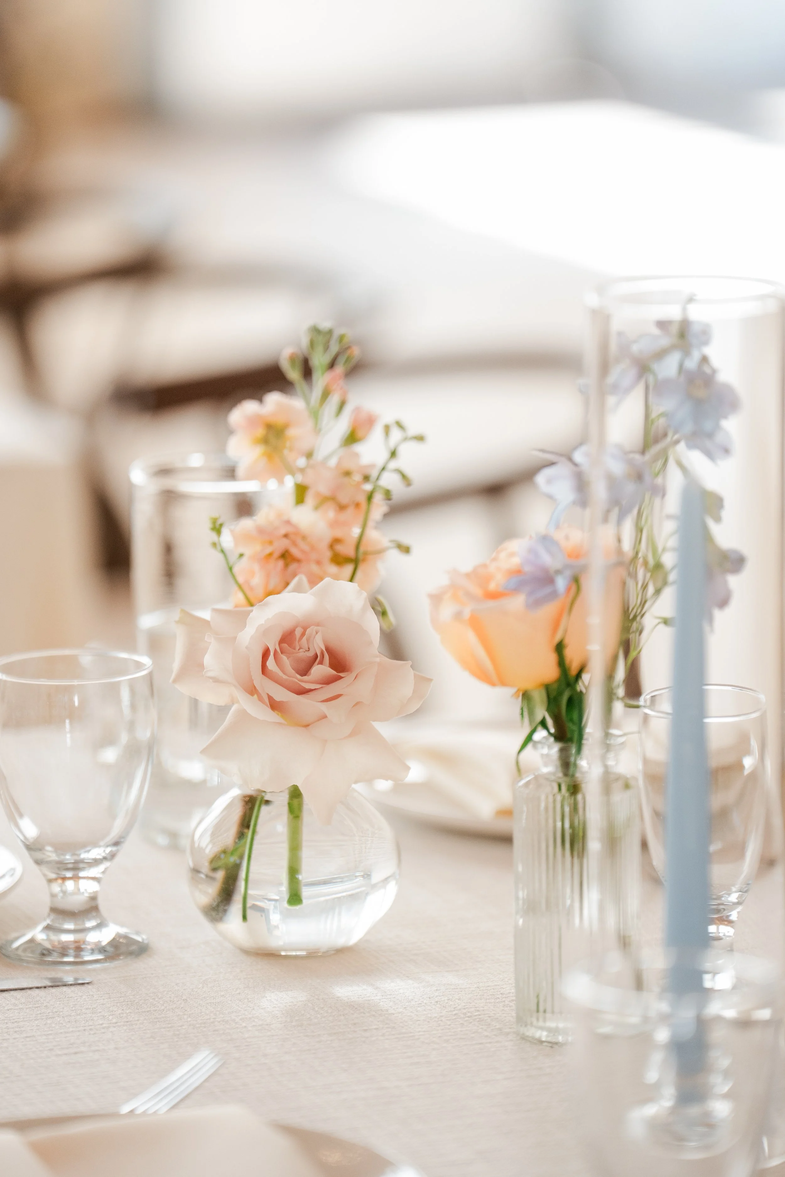 Close-up of a table centerpiece featuring pink and white flowers in glass vases, set on a white tablecloth with empty glasses around.
