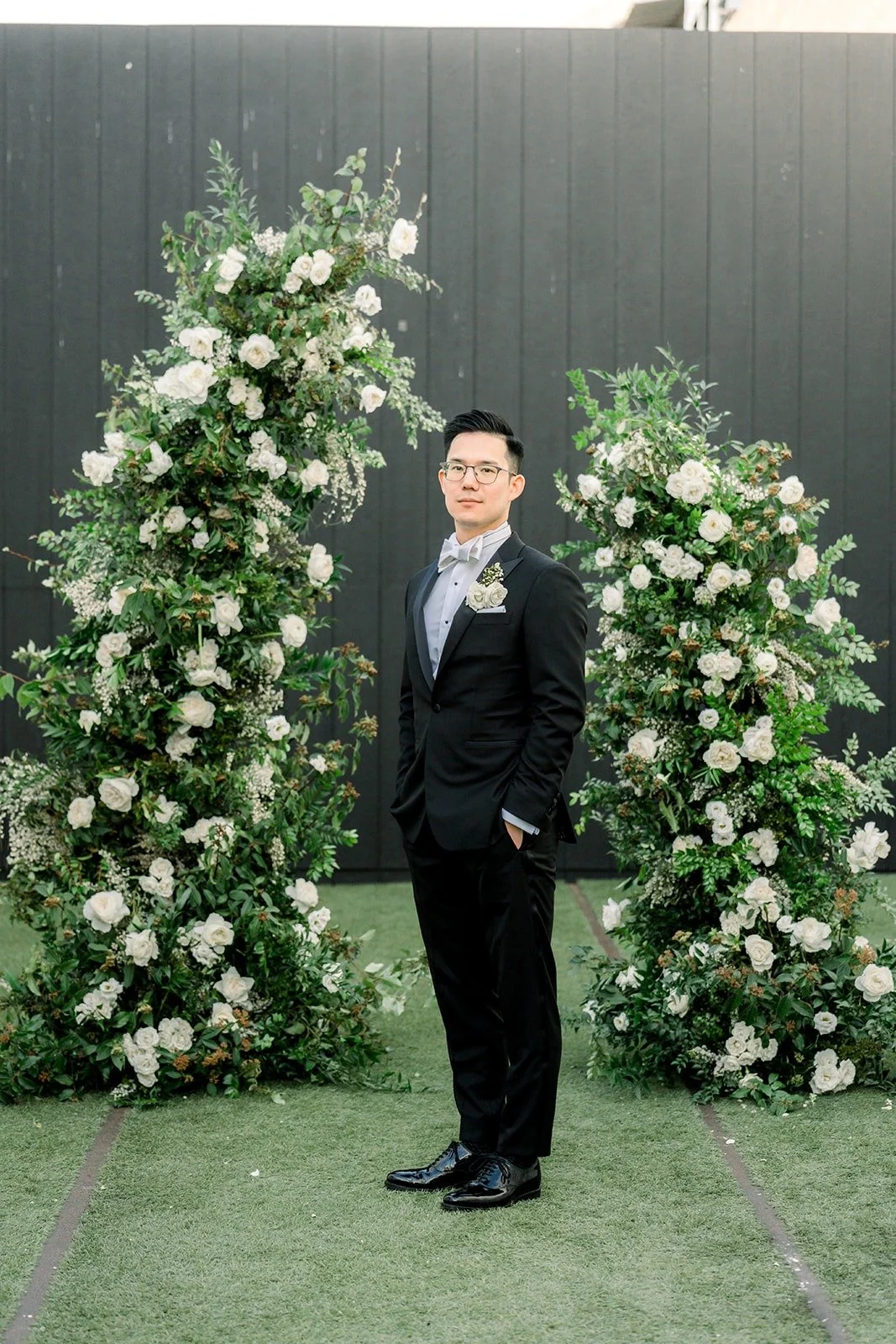 Alex stands between the white floral columns of the ceremony space, photographed by One and Only Production