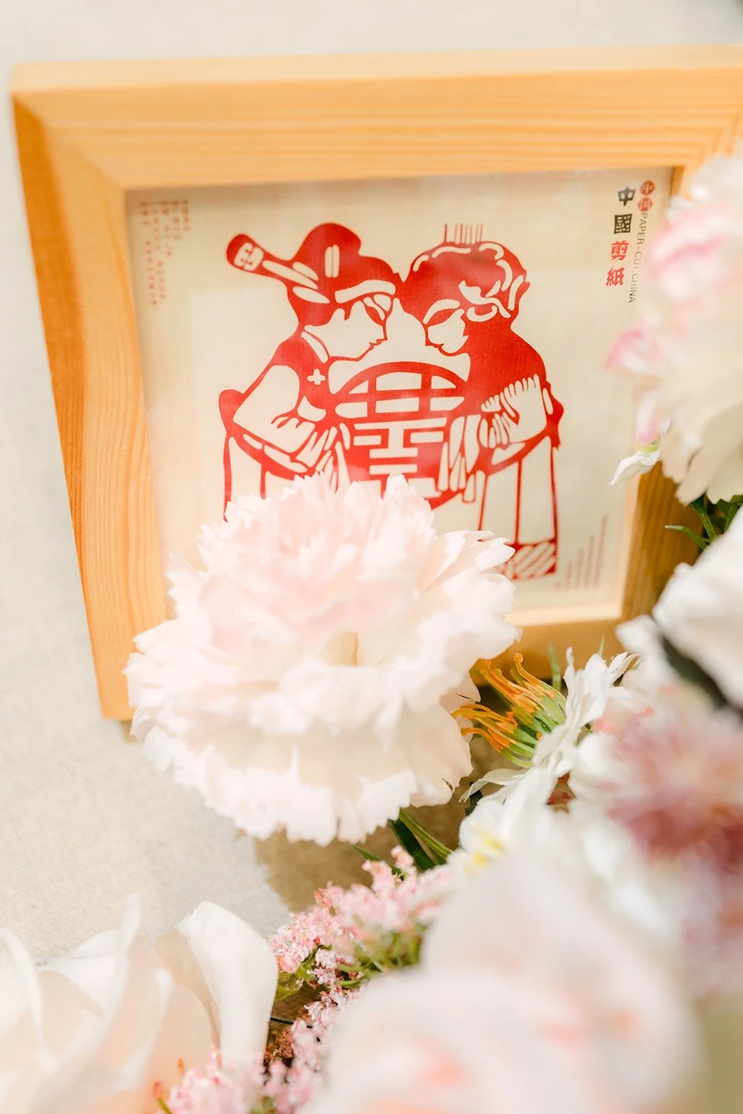A framed red paper cut of a double happiness couple surrounded by pink carnations, photographed by One and Only Production