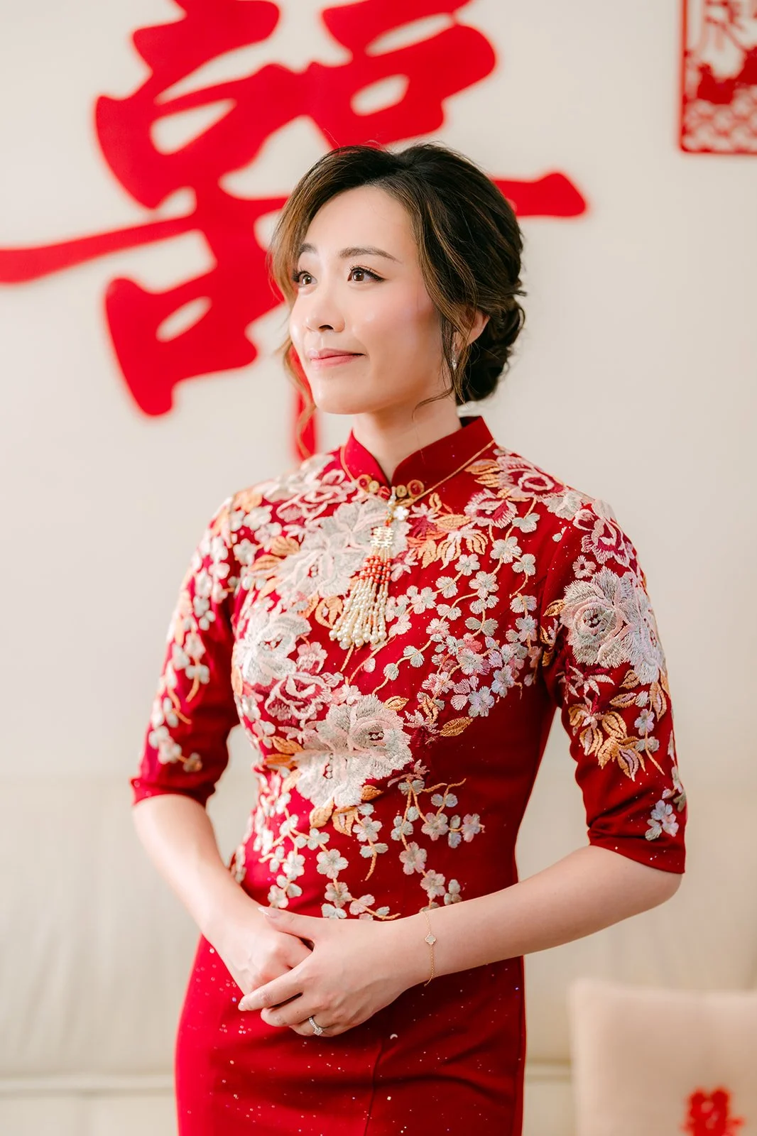 A bride in a red embroidered qipao in front of a double happiness papercut.