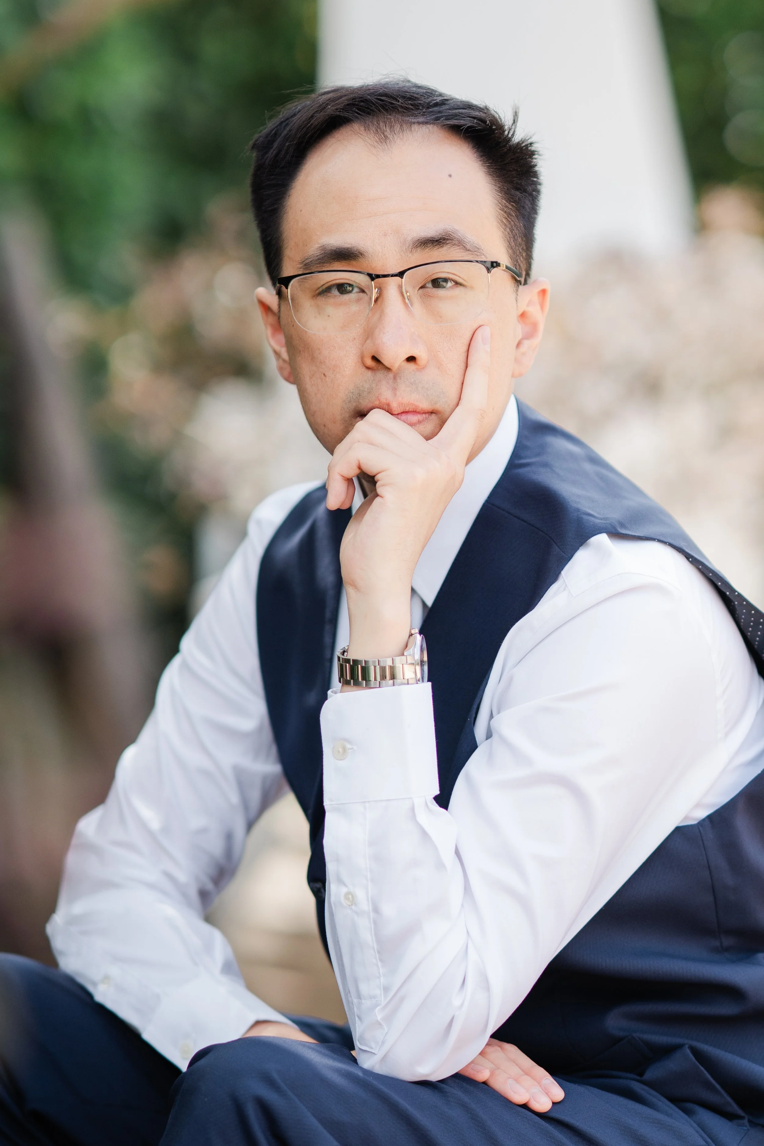 A man with glasses and dark hair, dressed in a white shirt and a dark vest, sitting outdoors with trees in the background. He is resting his chin on his hand and looking thoughtfully at the camera.