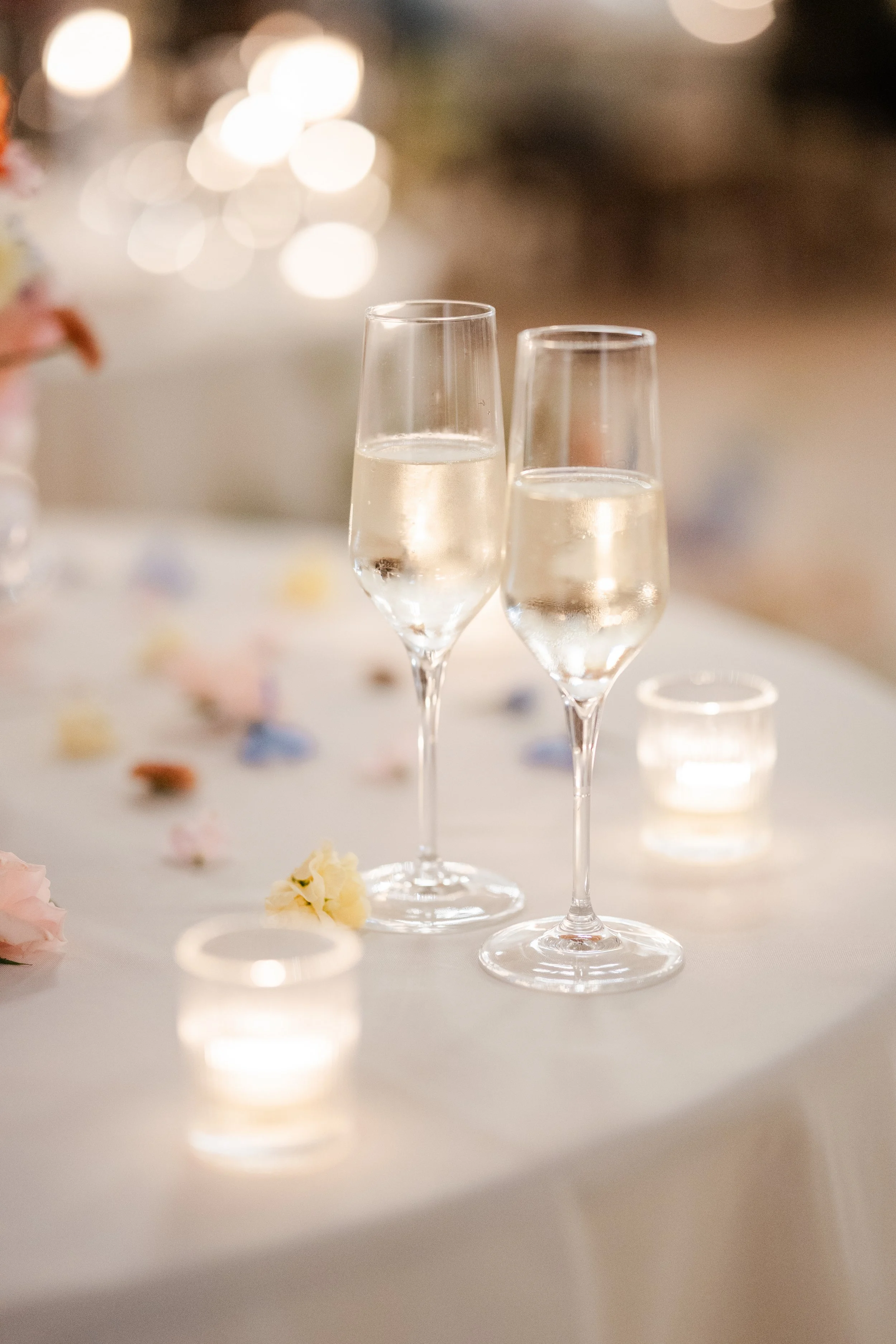 Two champagne flutes filled with sparkling wine or champagne on a decorated table with scattered flowers and a lit candle in the background.